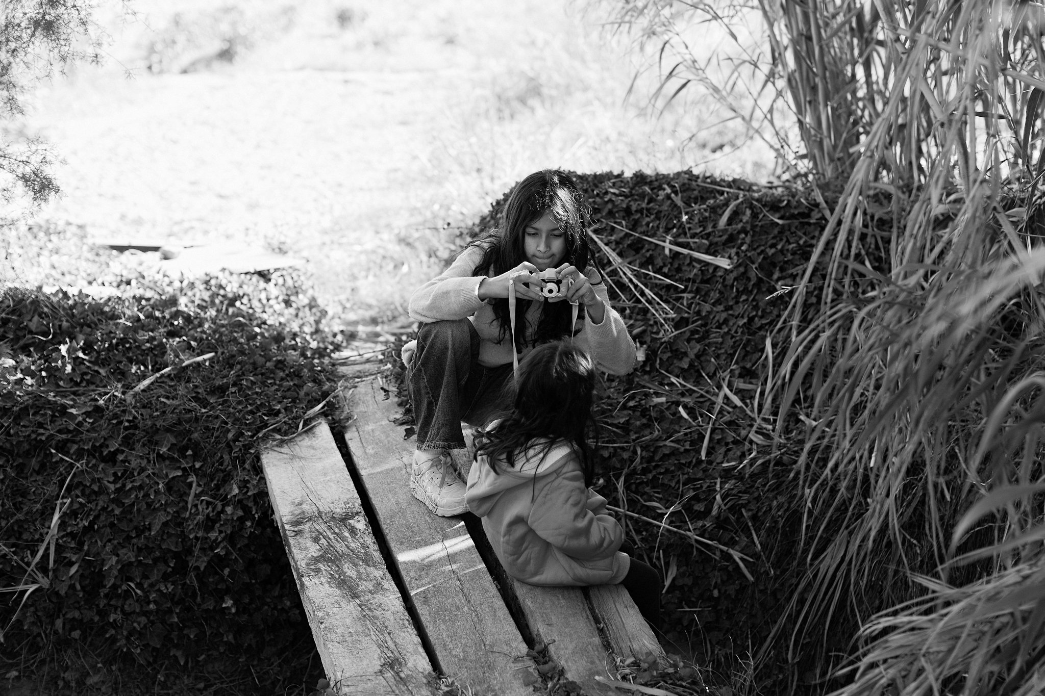 Una niña con cabello oscuro, vestida con una sudadera, está sentada en un tronco de árbol en un entorno natural rodeada de arbustos y maleza, sosteniendo una cámara fotográfica y mirando hacia ella.