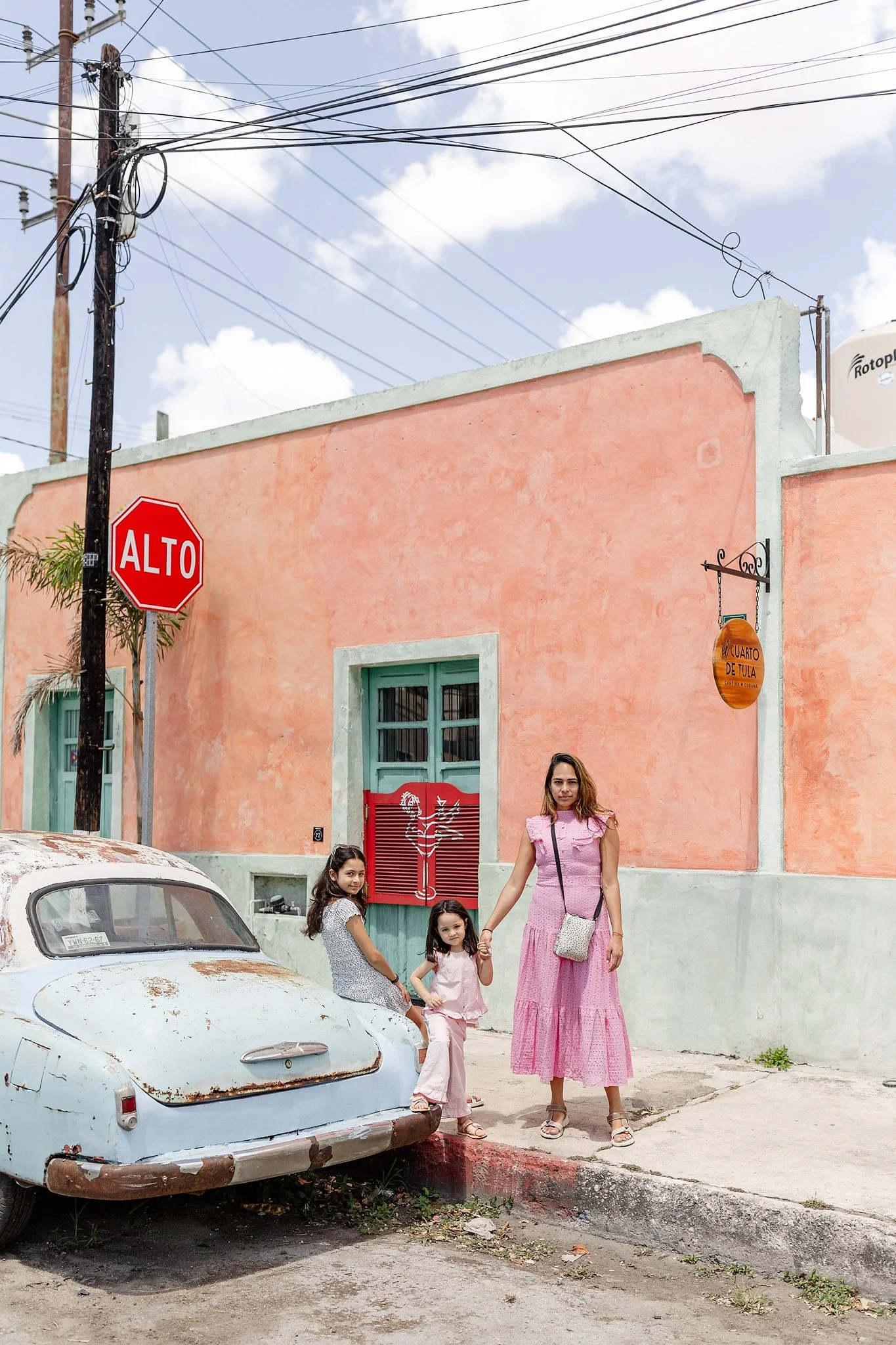 Mujer y dos niñas jóvenes sosteniendo las manos frente a una pared color durazno en una calle de ciudad, con un auto viejo y un señal de alto visible. La mujer lleva un vestido largo rosa y sandalias, las niñas ropa ligera y sandalias,  en un día soleado con cielo parcialmente nublado.