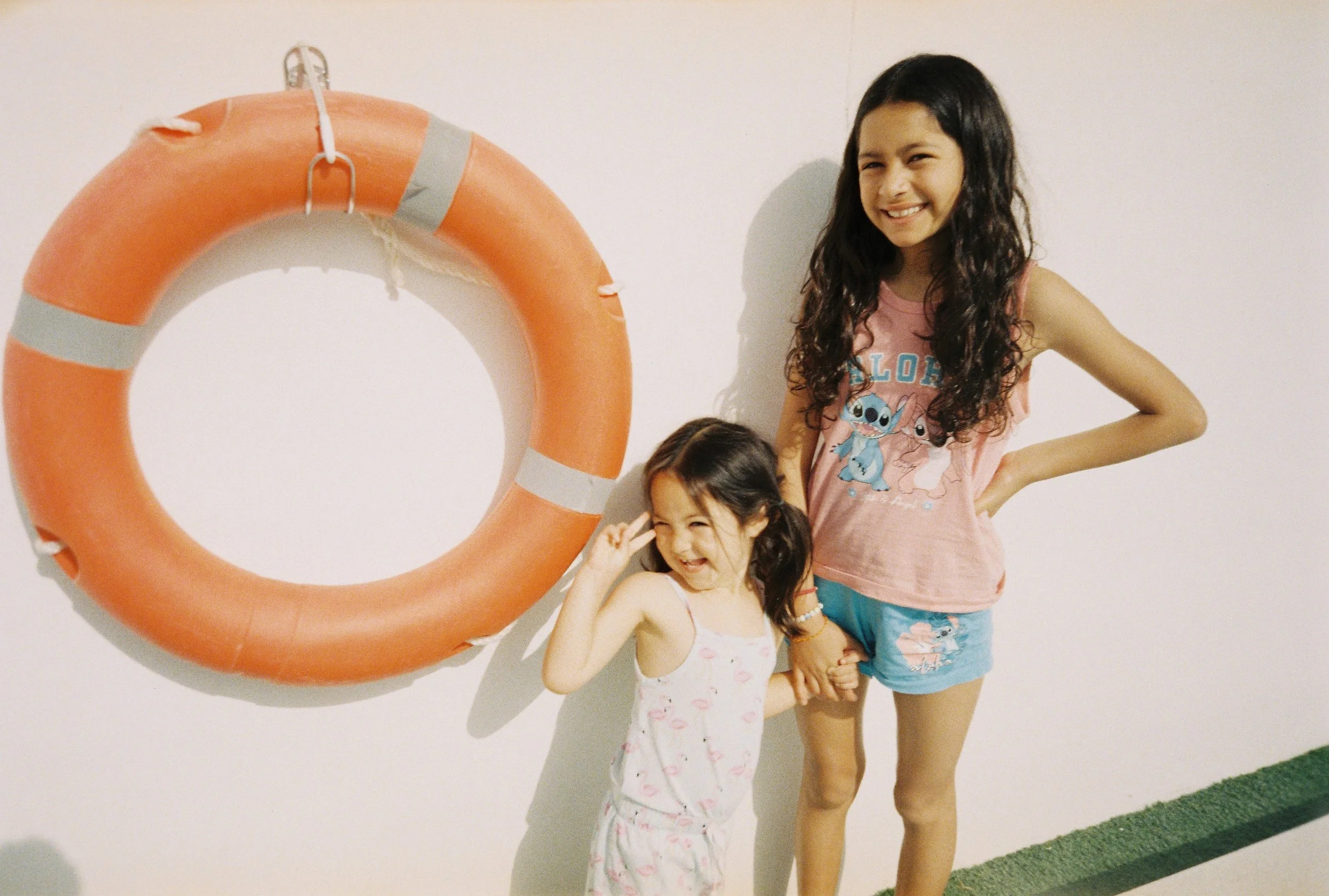 Dos niñas pequeñas y una joven sonriente posando junto a un salvavidas naranja en una pared blanca, con piso depastel y borde verde.