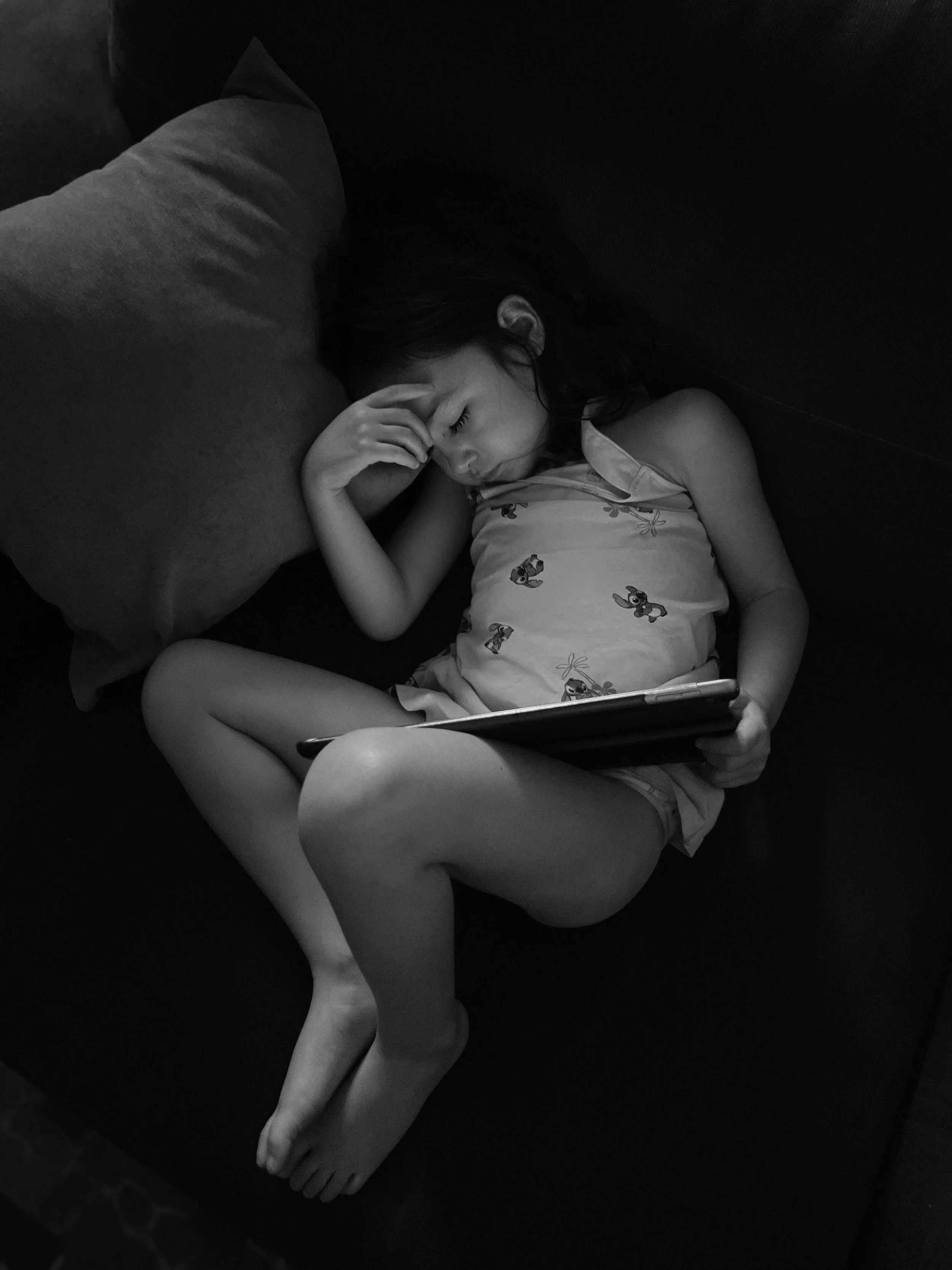 Una niña dormida en un sofá, usando un vestido con dibujos de perros, con las piernas dobladas y sosteniendo un teléfono móvil.