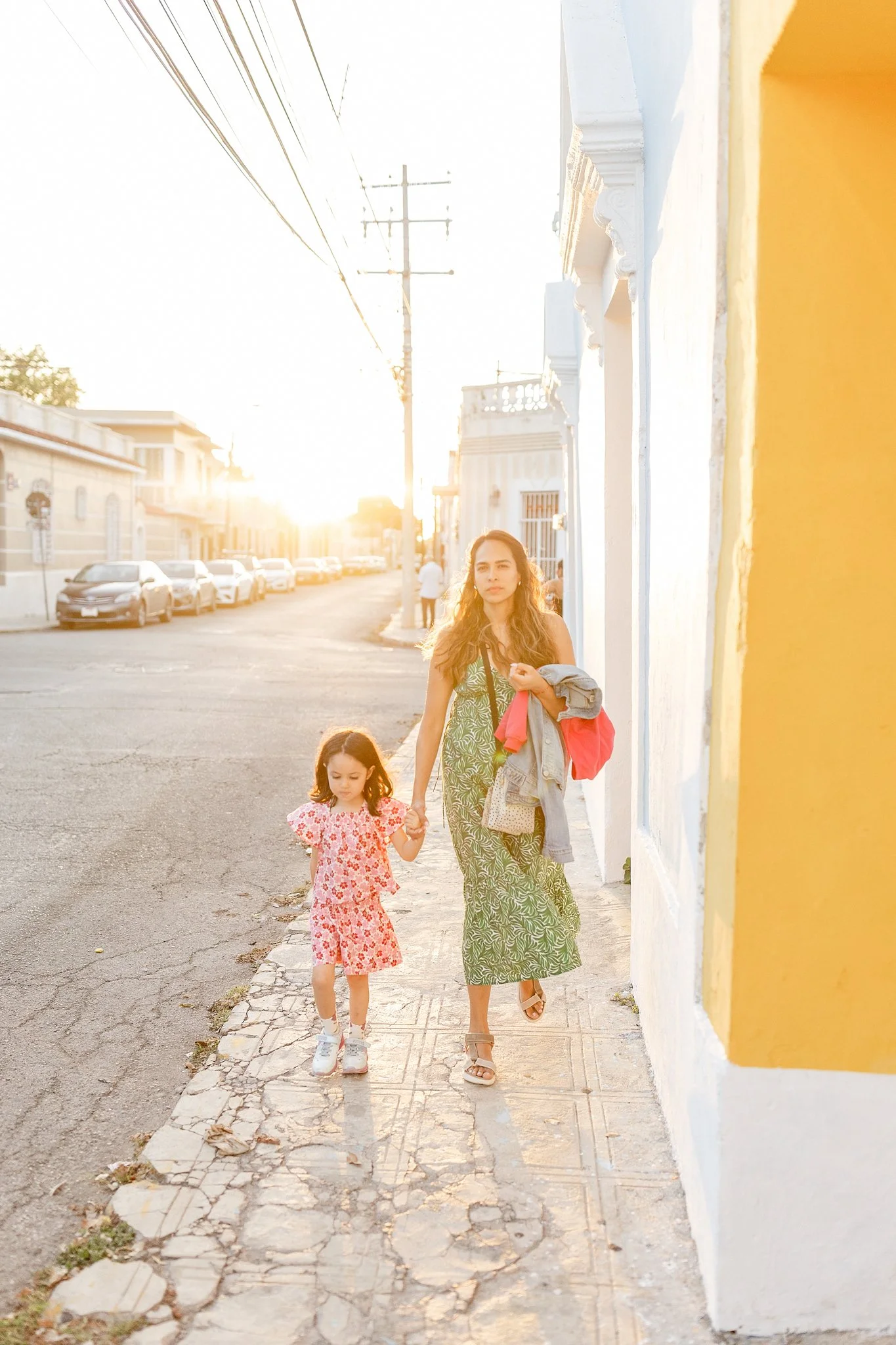 Mujer joven y niña caminando por la acera en una calle soleada, con casas coloridas y coches estacionados.