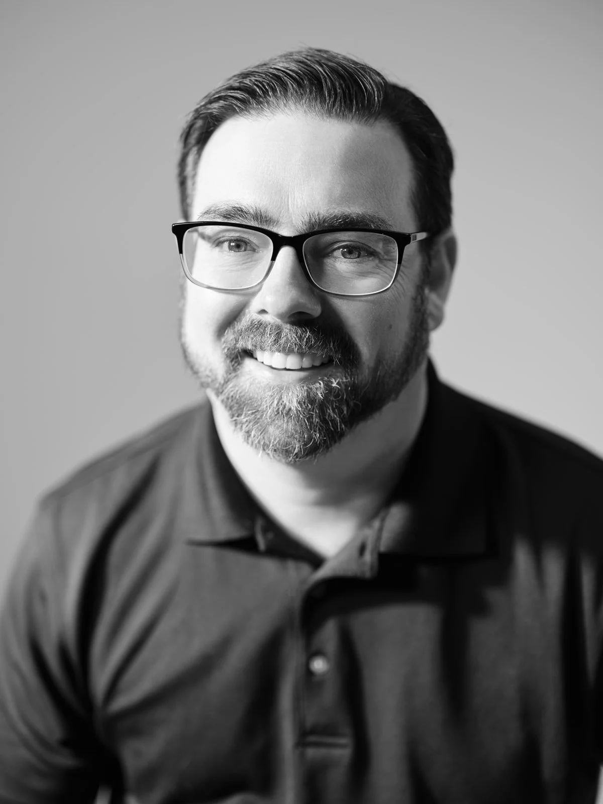 Riverbend Appraisals - JOSH AMENT - A man with glasses, brown hair, and a beard wearing a dark suit and white shirt, smiling at the camera.