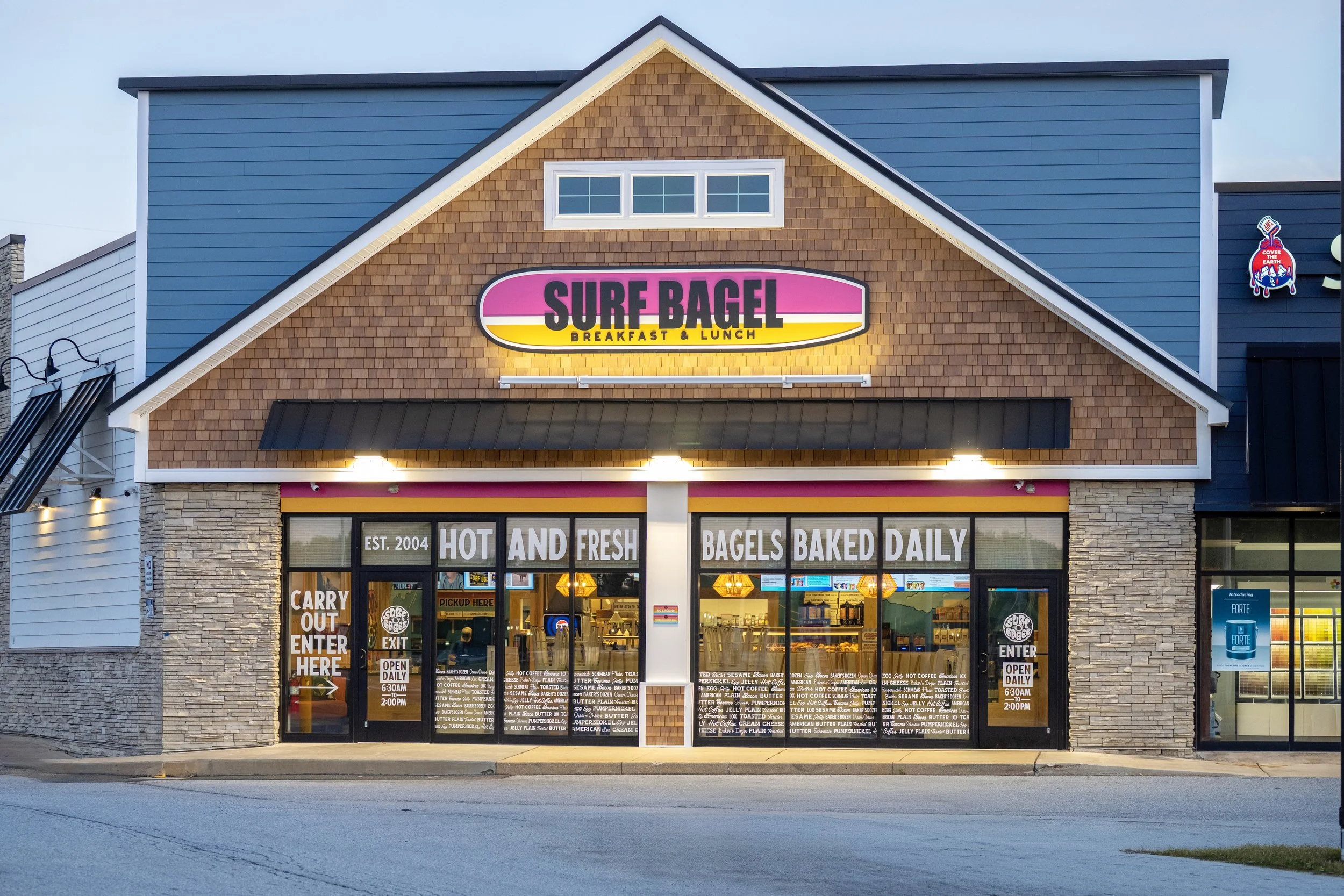 The storefront of Surf Bagel, a breakfast and lunch restaurant, with a large sign displaying its name. The building features a rustic brick facade with a black awning and large glass doors. Inside, the menu is visible in white lettering on the doors, advertising hot and fresh bagels baked daily, carry-out options, and open hours from 6:30 AM to 2:00 PM.
