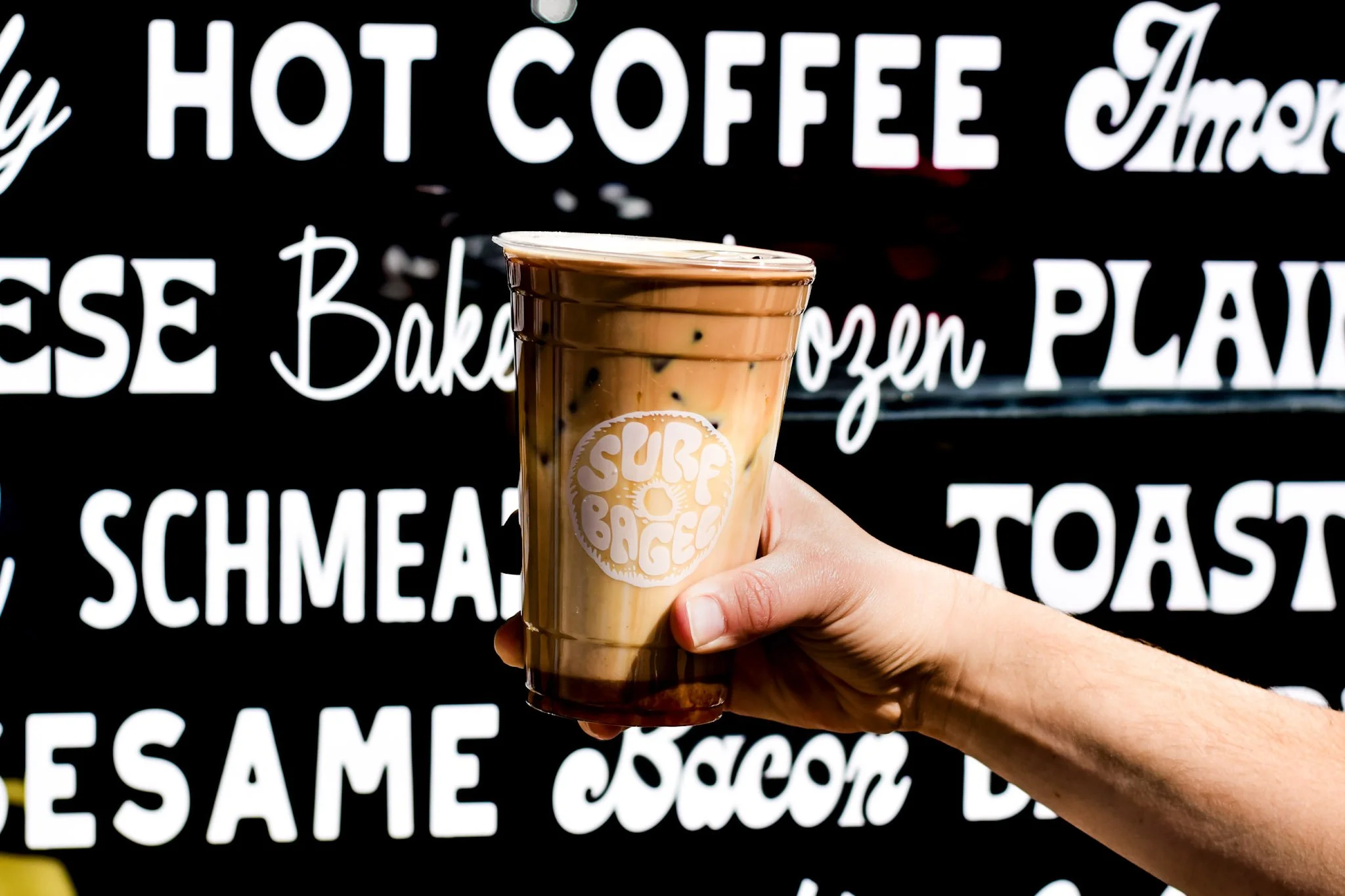 Hand holding a Surf Bagel Caramel Macchiato in front of the Surf Bagel Storefront