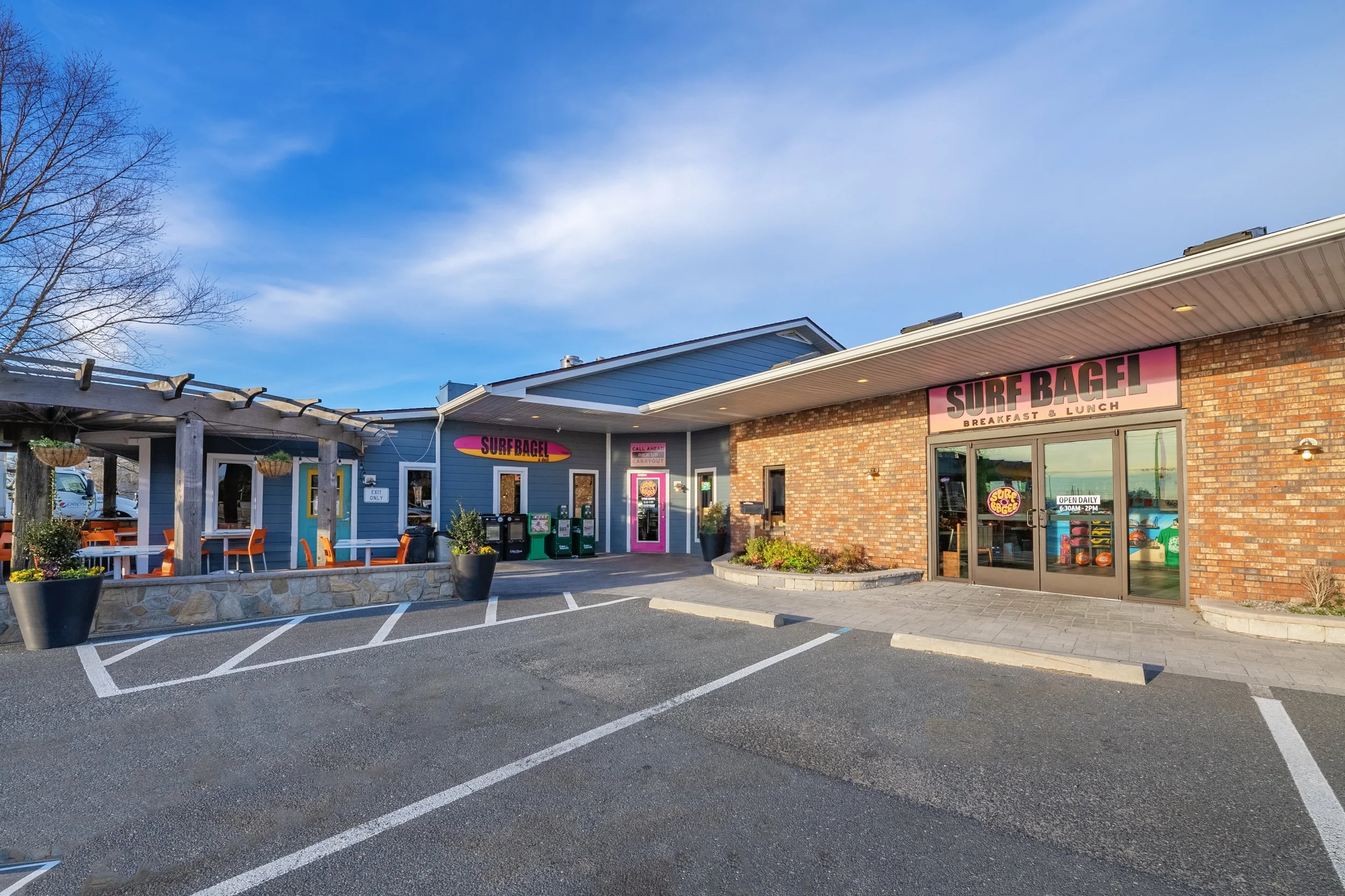 Exterior view of a restaurant called Surf Bagel with outdoor seating, a parking lot, and a clear blue sky.