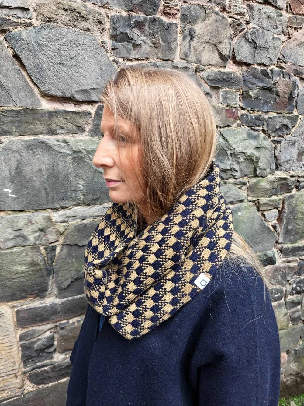 Lady with long hair wearing an Archipelago soft wool grey patterned snood in front of a stone wall.