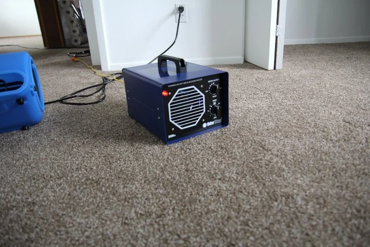 A portable space heater sitting on a beige carpeted floor near an open door, with a white wall and some cords in the background.