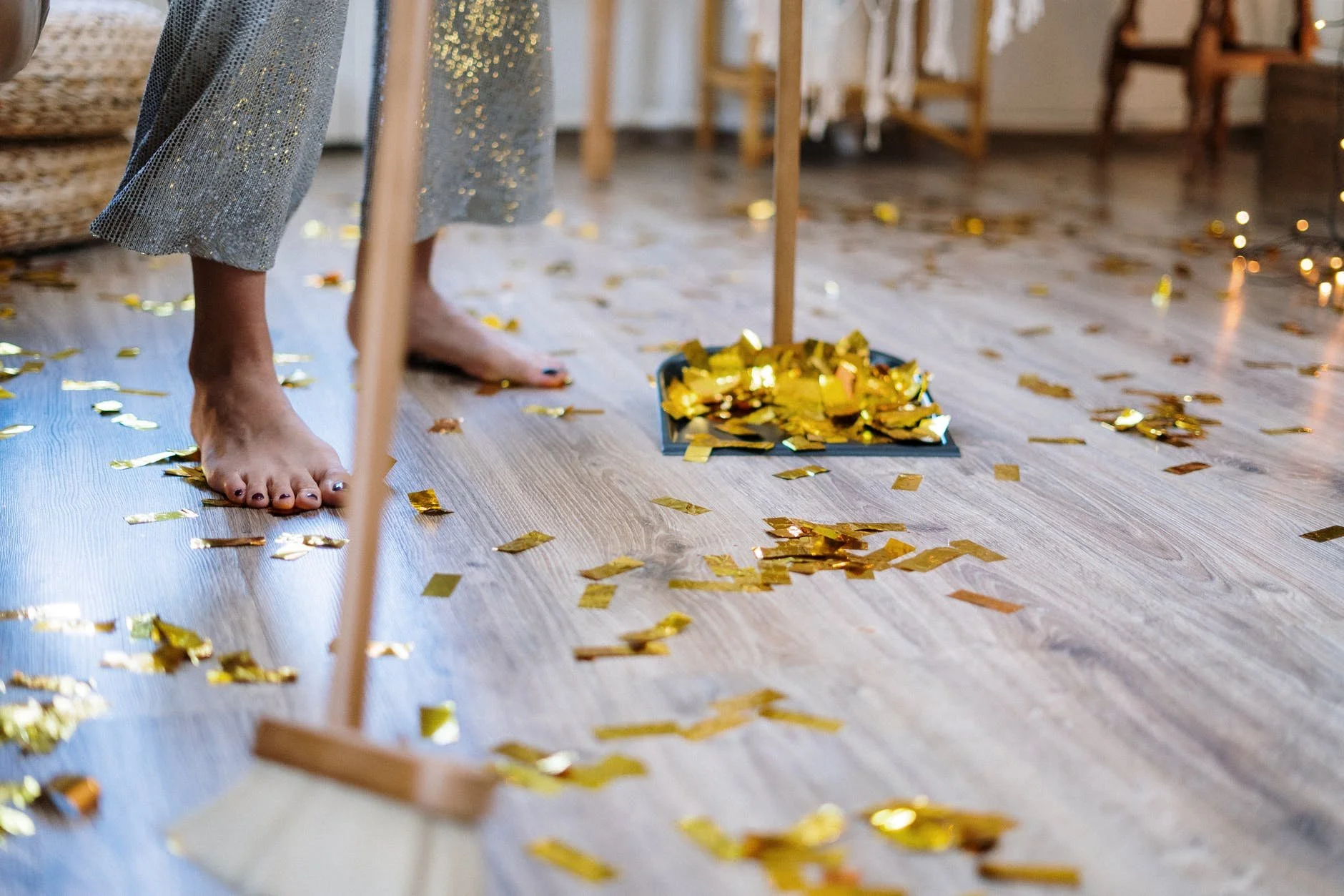 Bare feet on wooden floor with gold confetti scattered around and a dustpan filled with gold confetti in the background.