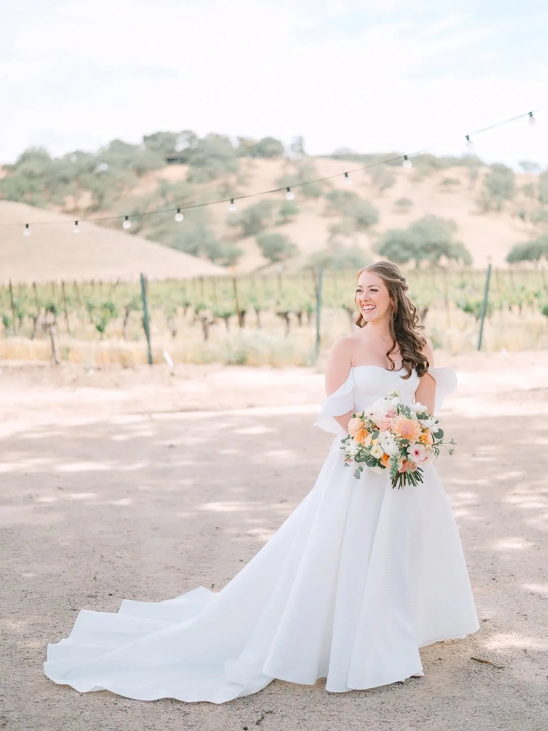 Bride looking left while holding peach and pink bouquet in front of vines at luxury California vineyard venue Cass Winery in Paso Robles.