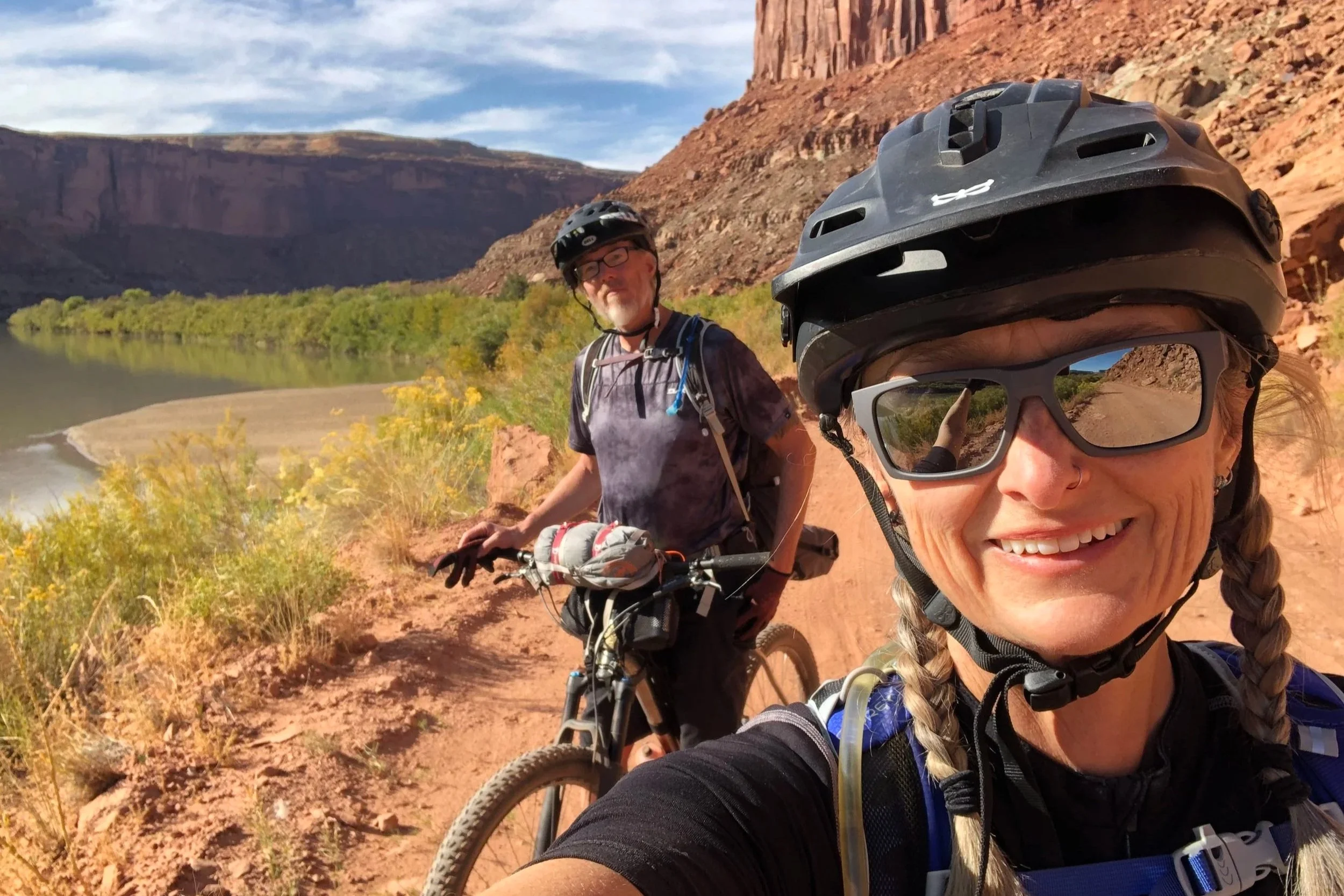 Kevin and Michelle on their bikes at the White Rim Trail bikepacking in Utah. Red rock cliffs and a river in the background.