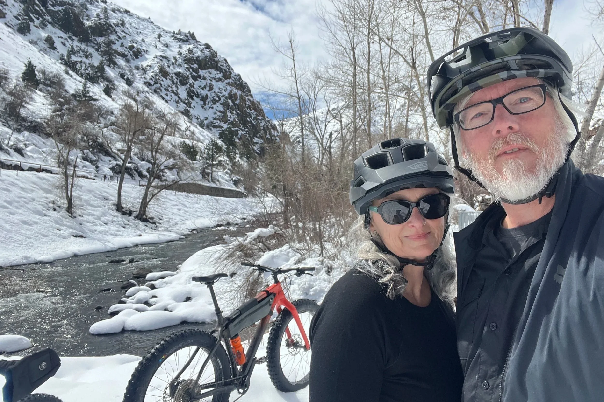 Kevin and Michelle fatbiking in Golden along Clear Creek with the creek and snowy banks in the background.