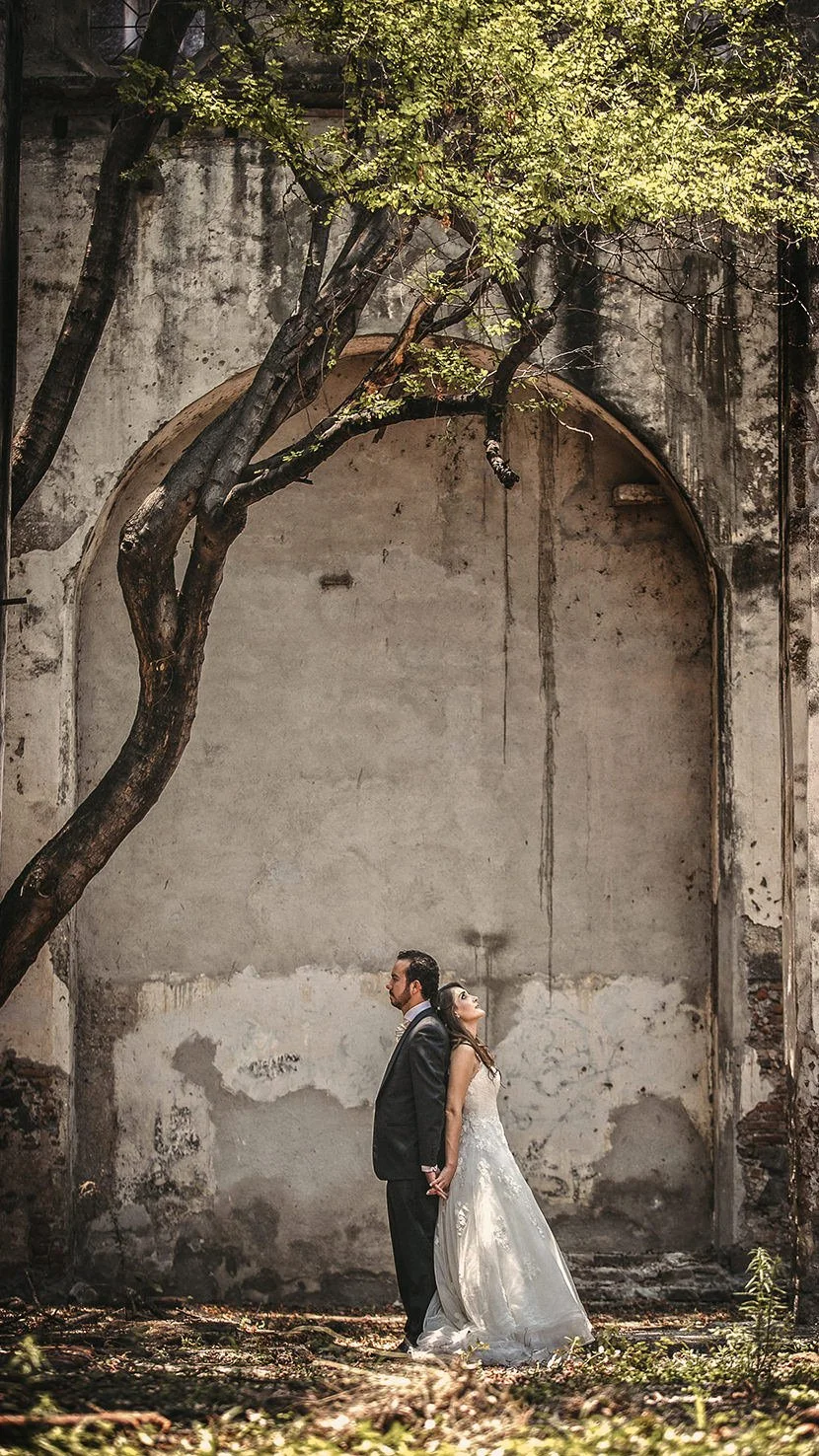 A bride and groom holding hands, standing back to back outdoors in front of a weathered wall with a large arch and a tree with green leaves overhead.