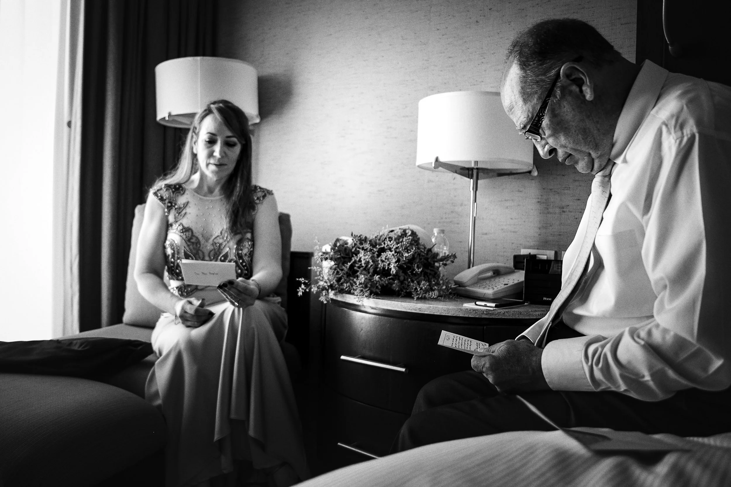 Fotografía en blanco y negro de madre de la novia sentada durante preparación, acompañada por el padre de la novia en interior.