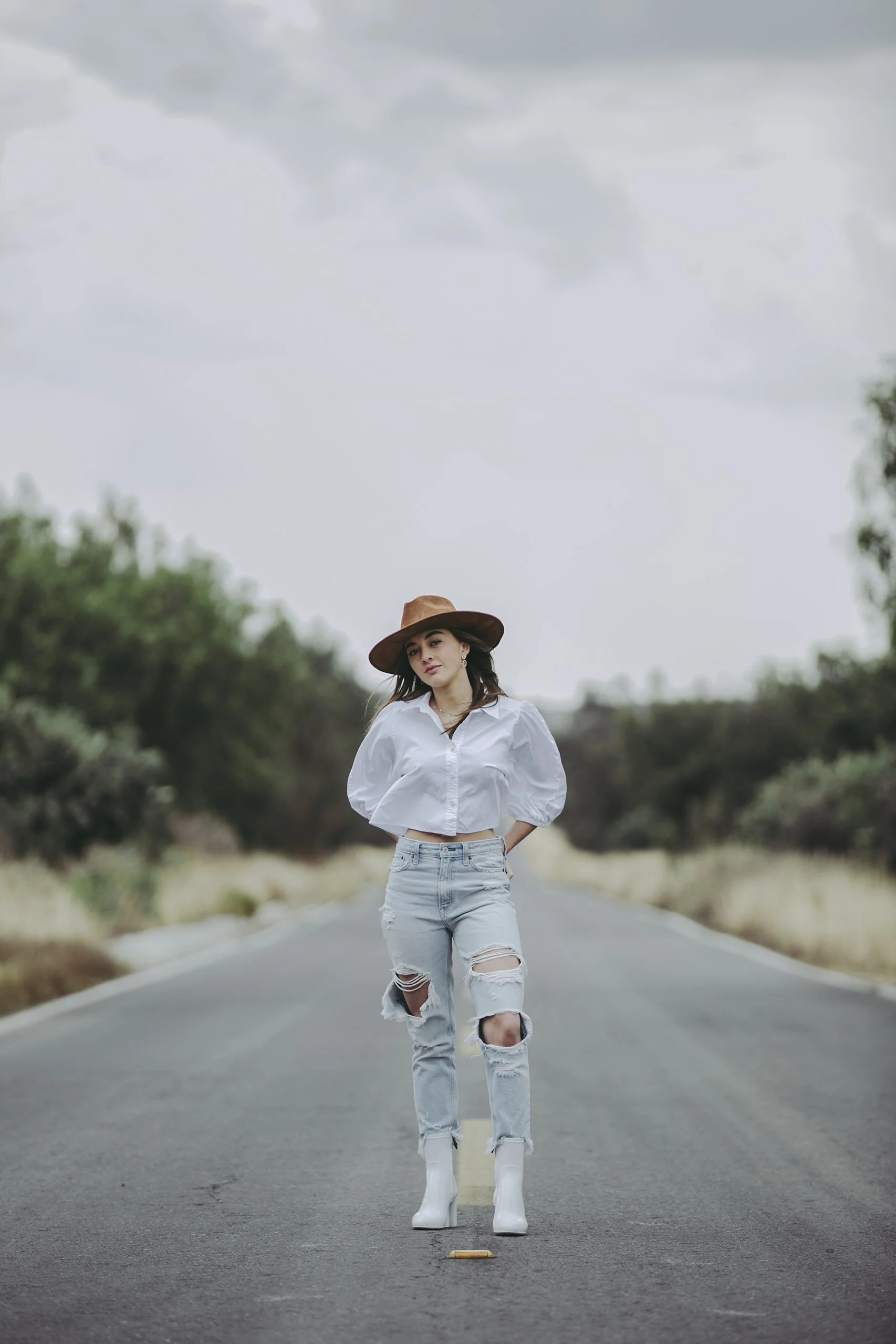 Retrato de mujer caminando por carretera rural, vestimenta clara y sombrero, mirada directa.