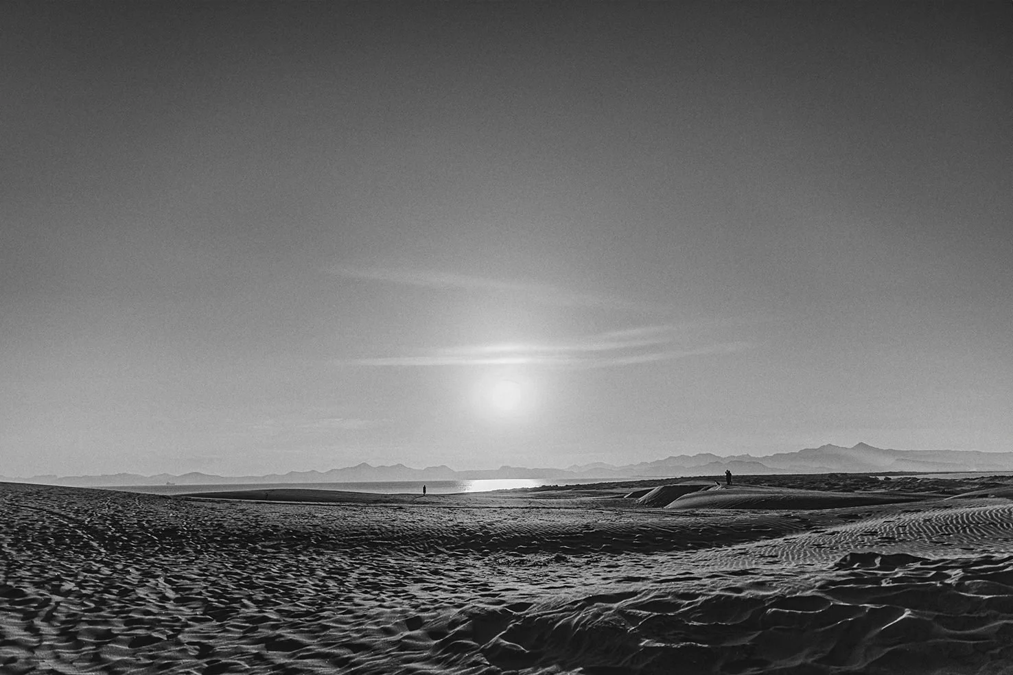 Black and white photo of a desert landscape with sand dunes, a calm body of water, distant mountains, and a setting or rising sun in the sky. Two small figures are visible in the distance.