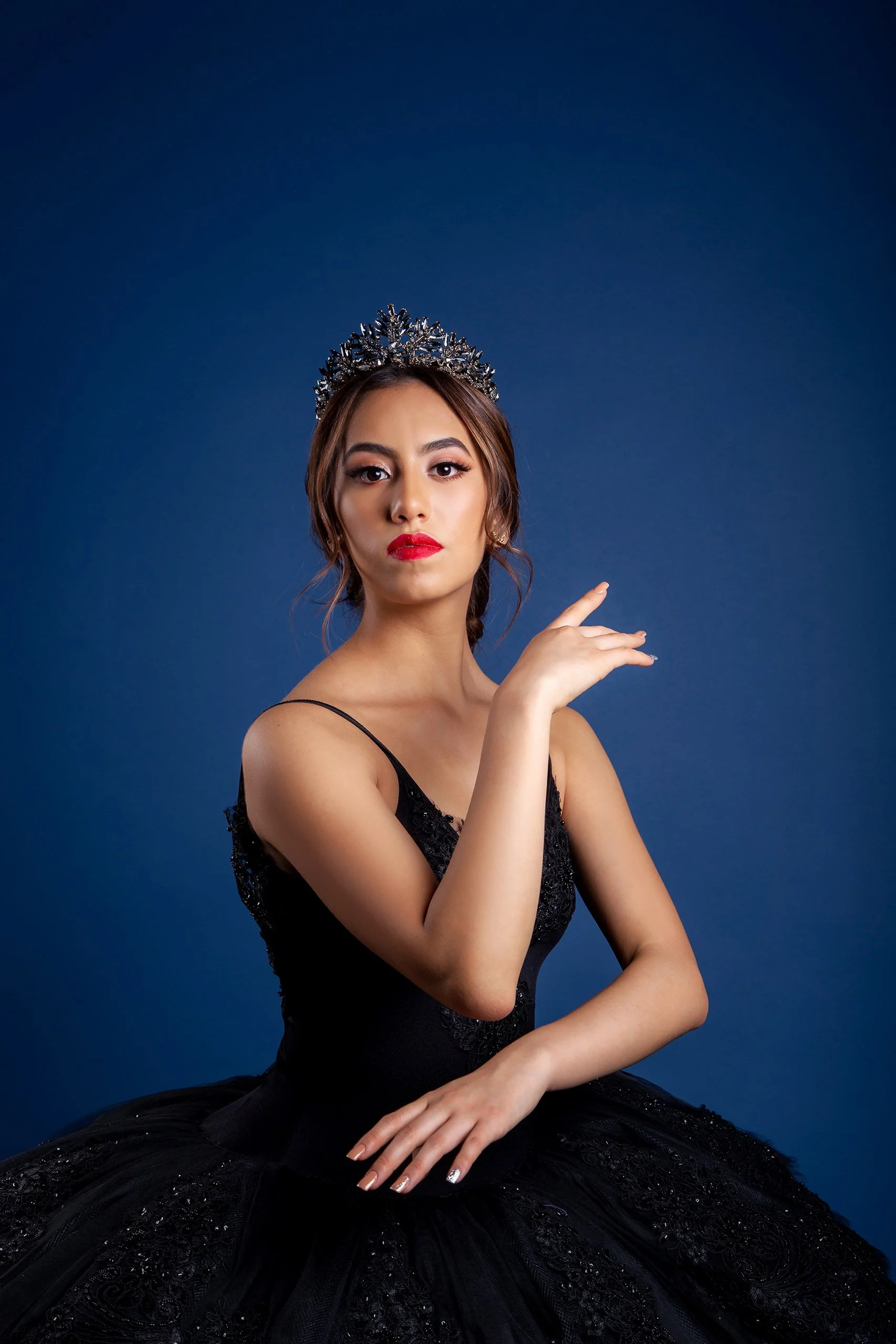 Retrato de mujer en estudio con vestido negro y tiara, fondo azul y gesto elegante mirando a cámara.