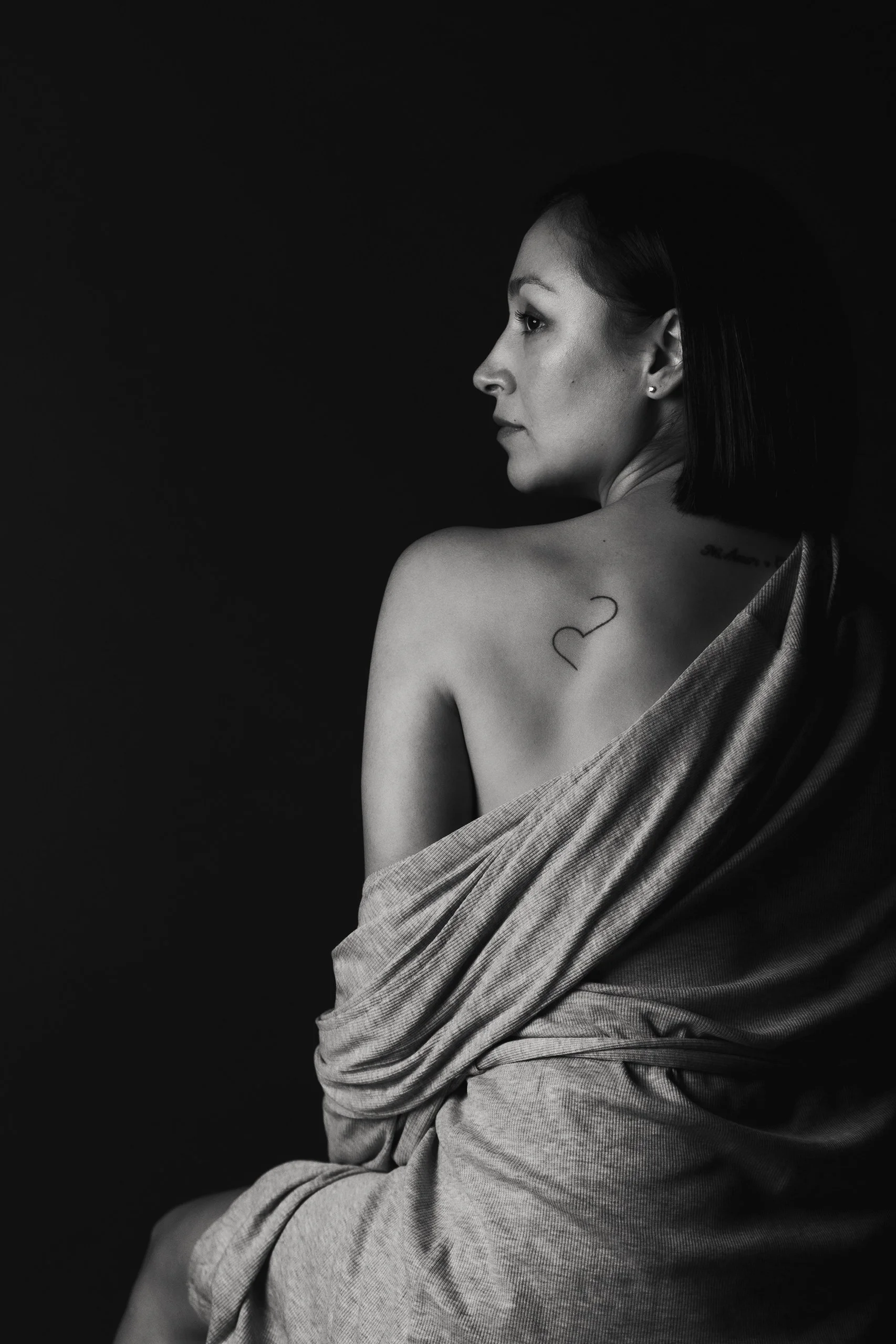 Retrato en blanco y negro de mujer de espaldas, hombros descubiertos y tatuaje visible en la espalda.