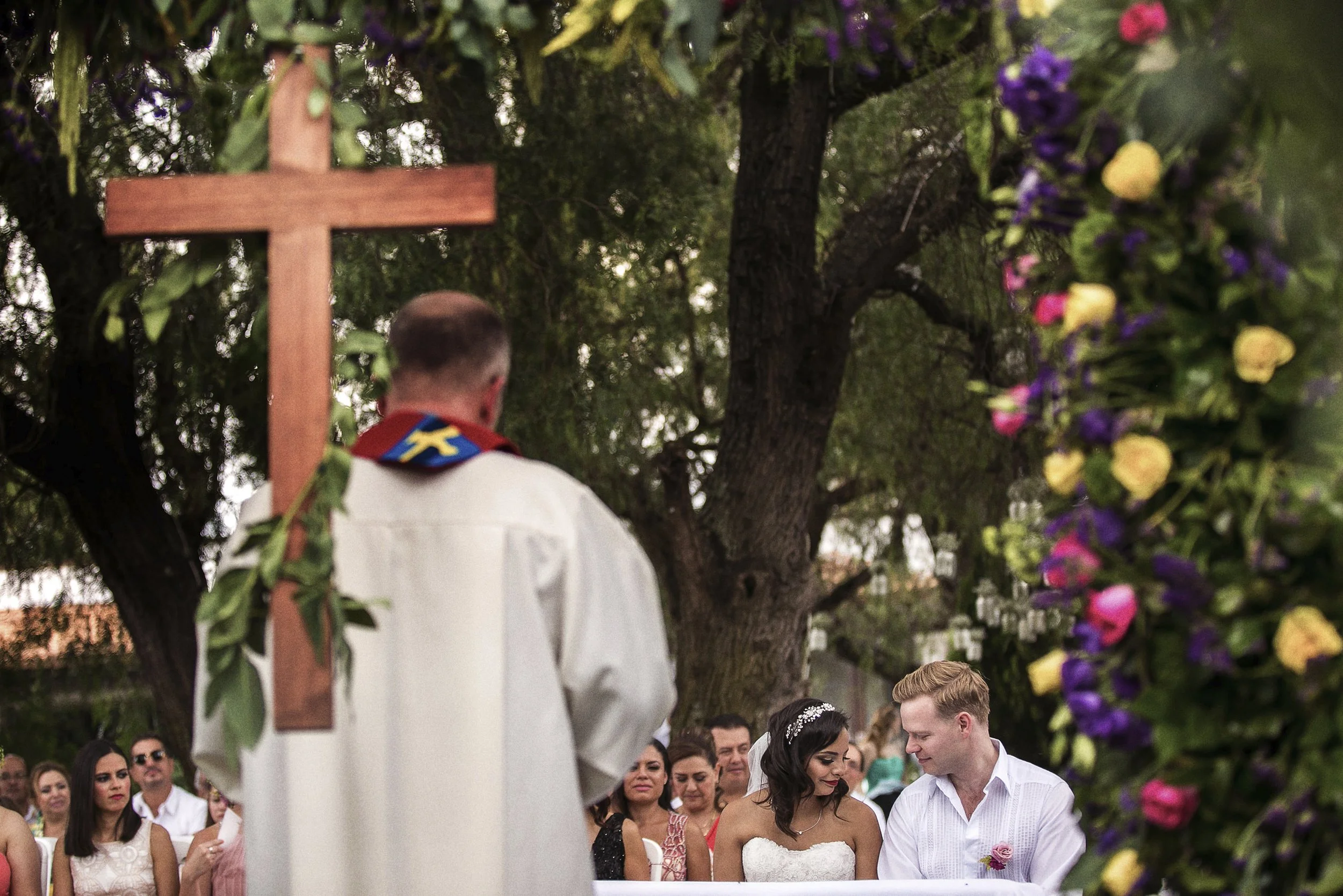Fotografía documental de ceremonia religiosa al aire libre, sacerdote y novios frente a invitados.