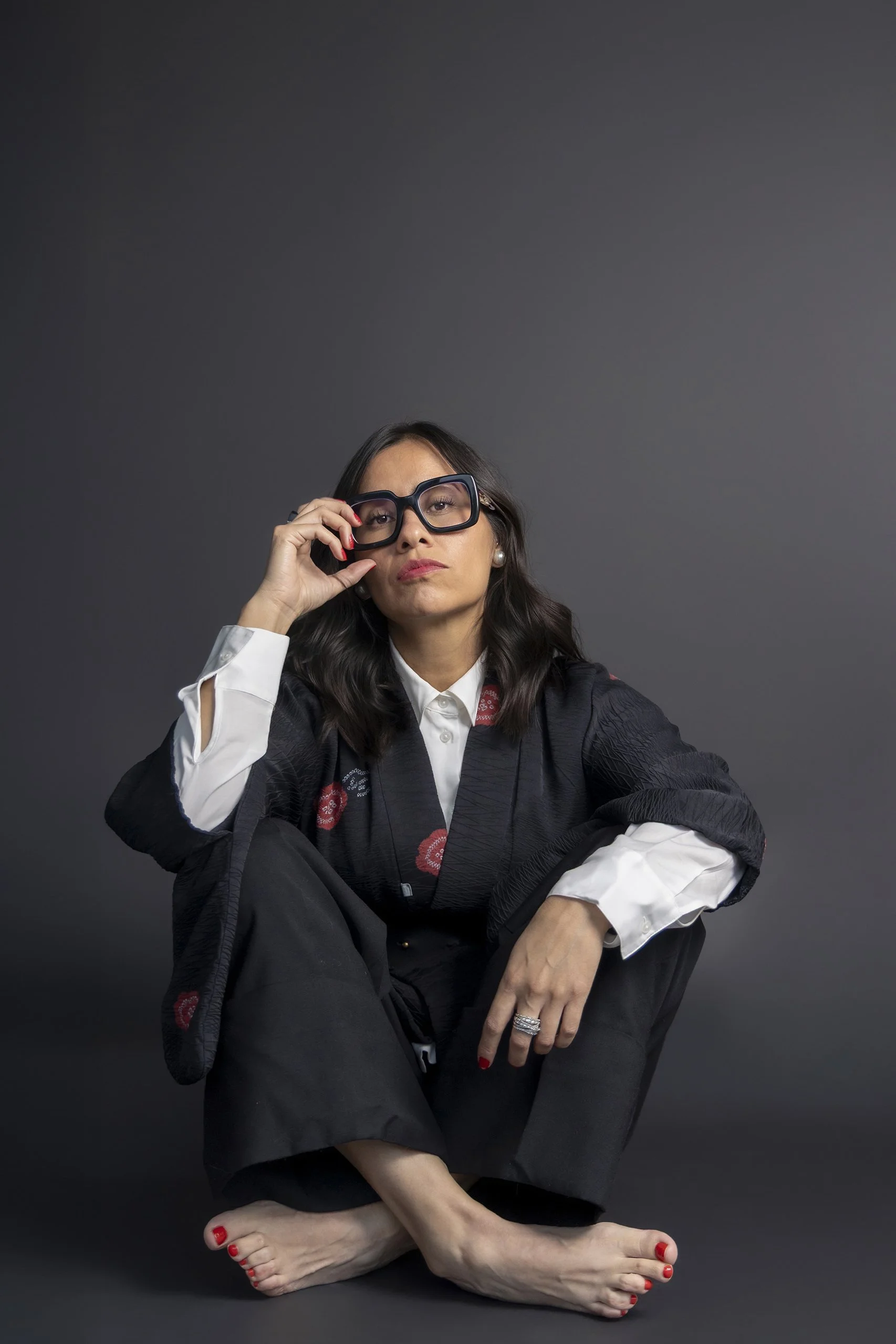 Retrato de mujer sentada en estudio, traje oscuro, lentes y gesto relajado mirando hacia arriba.
