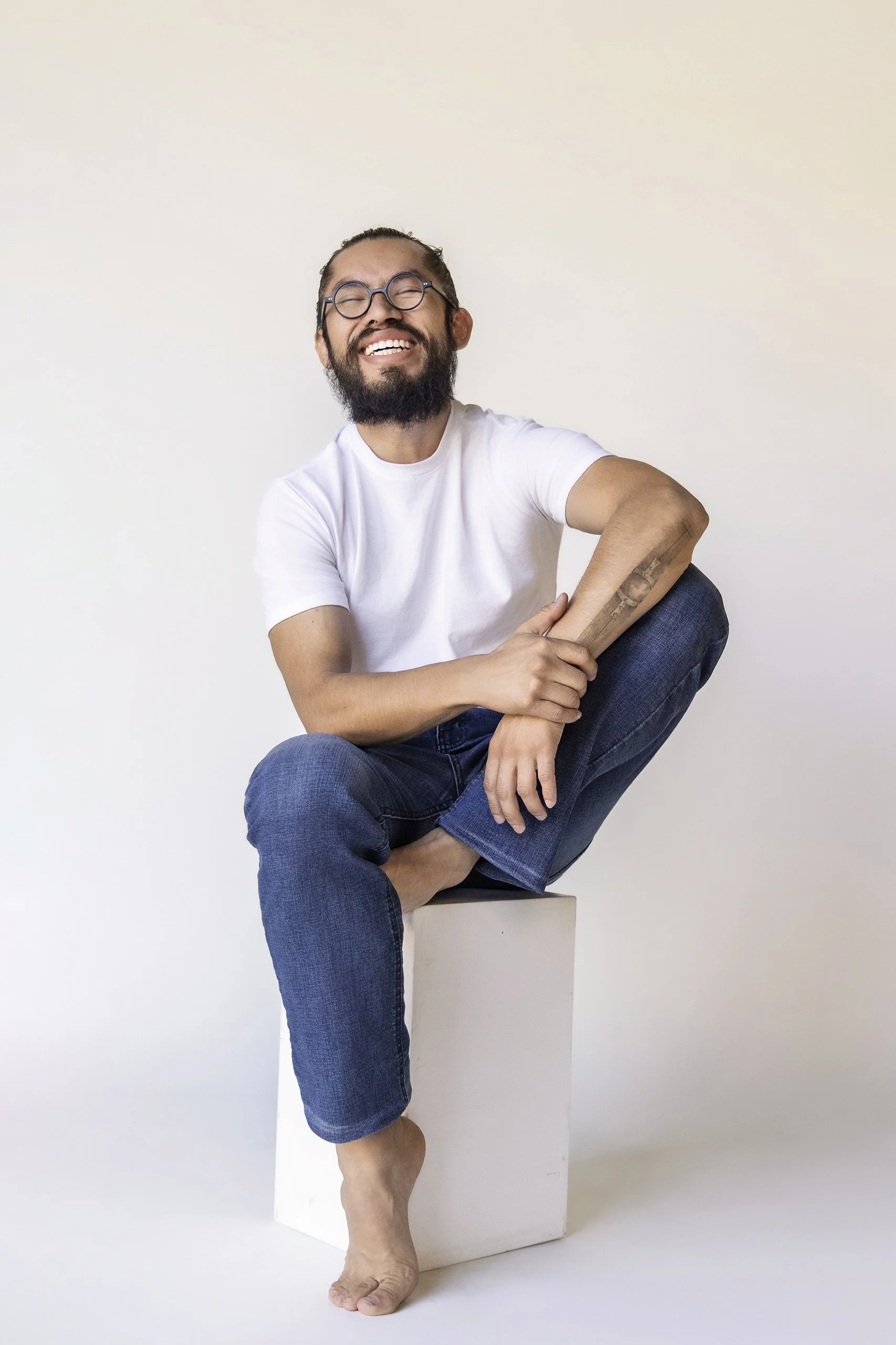 Retrato de hombre sentado con postura relajada y sonrisa abierta, fondo claro y luz natural.