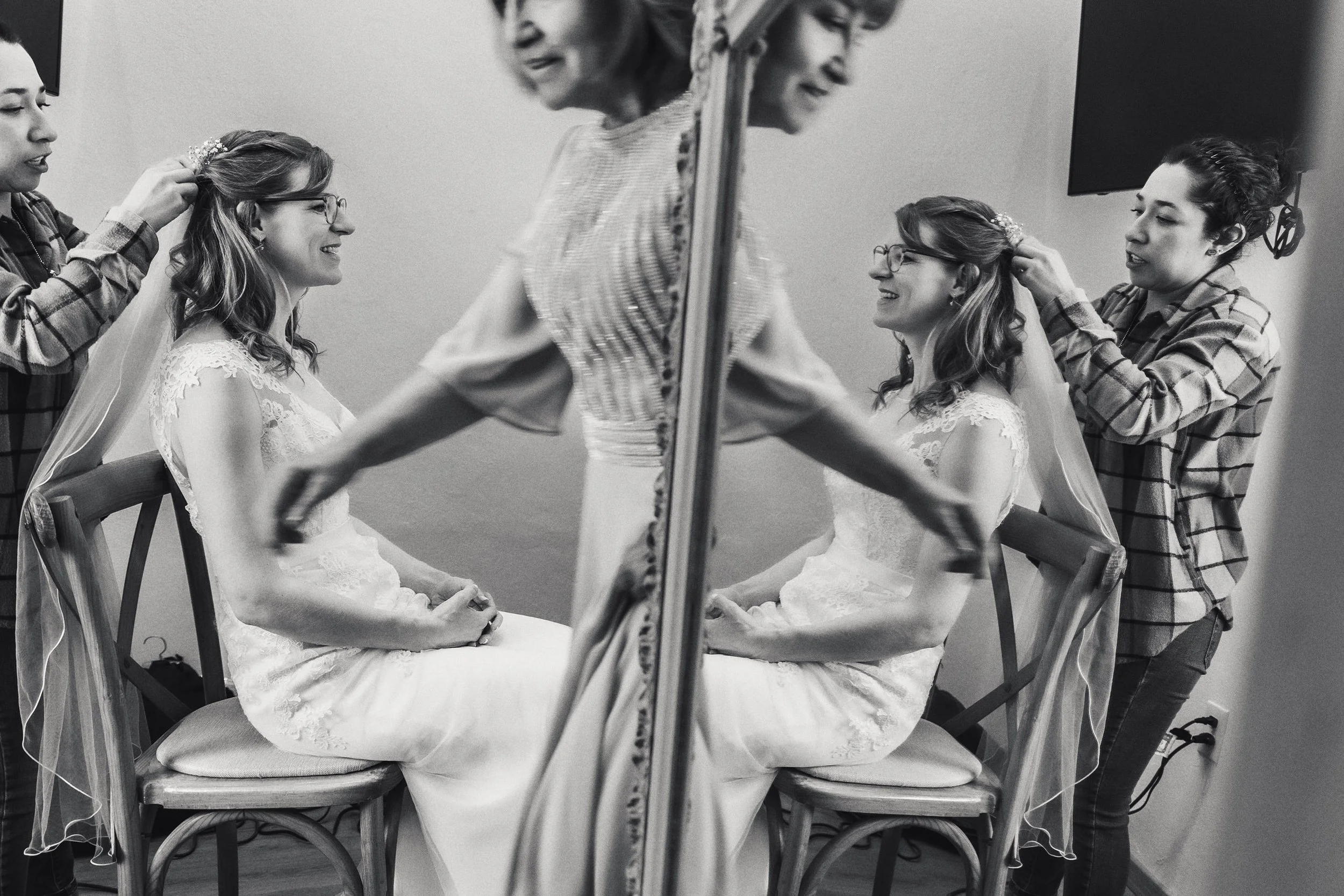 Fotografía en blanco y negro de novia sentada frente a espejo, ajuste de vestido y gesto íntimo.