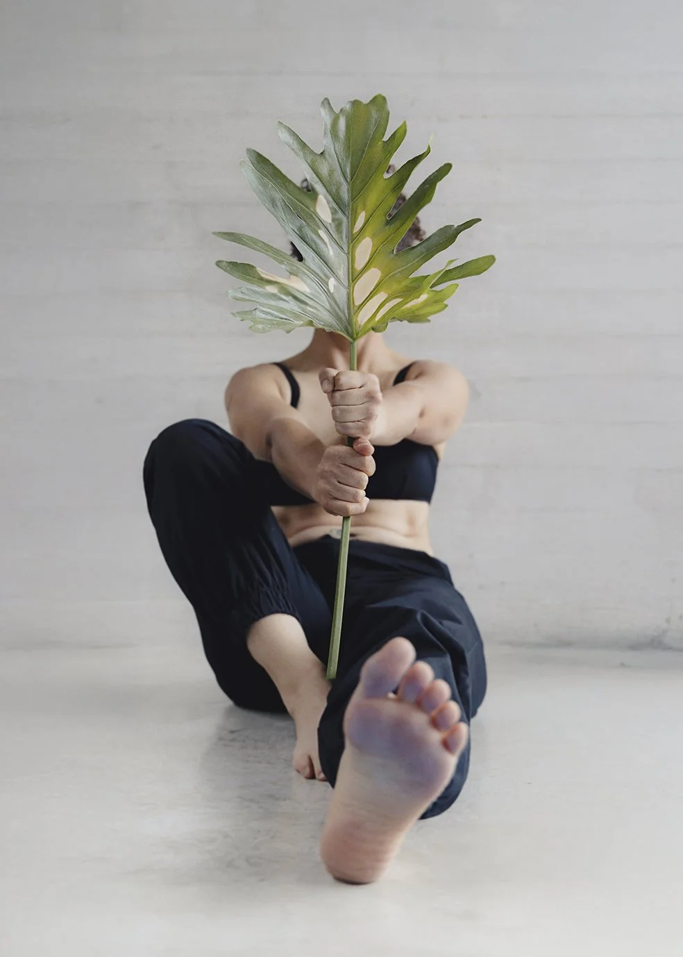 Person sitting on the floor, holding a large green leaf in front of their face, with their foot extended towards the camera.