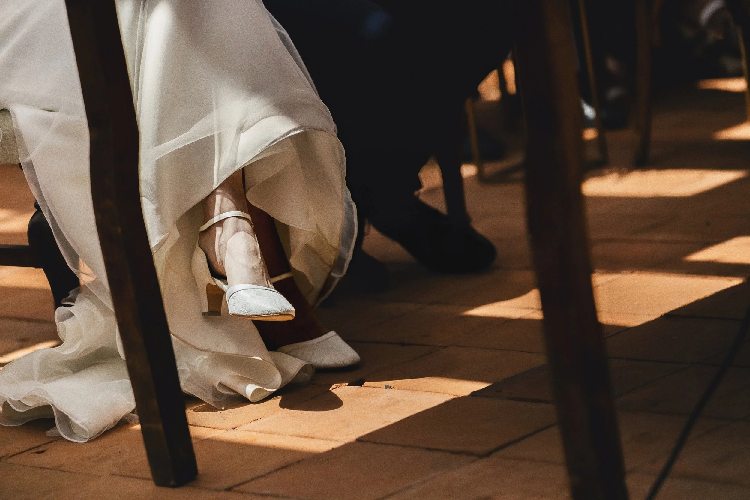 Detalle de zapatos de novia bajo mesa durante la recepción, luz cálida y encuadre bajo.