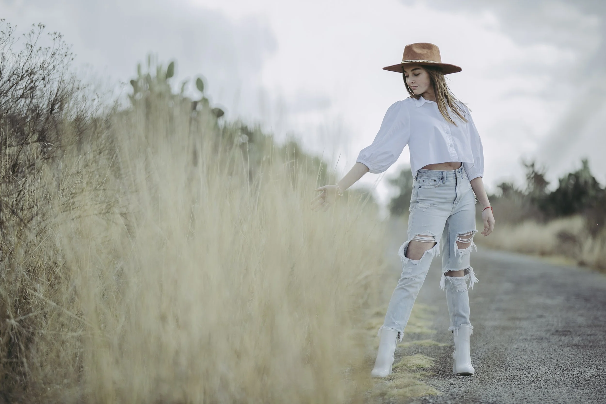 Retrato de mujer caminando entre vegetación alta, vestimenta clara y gesto contemplativo.