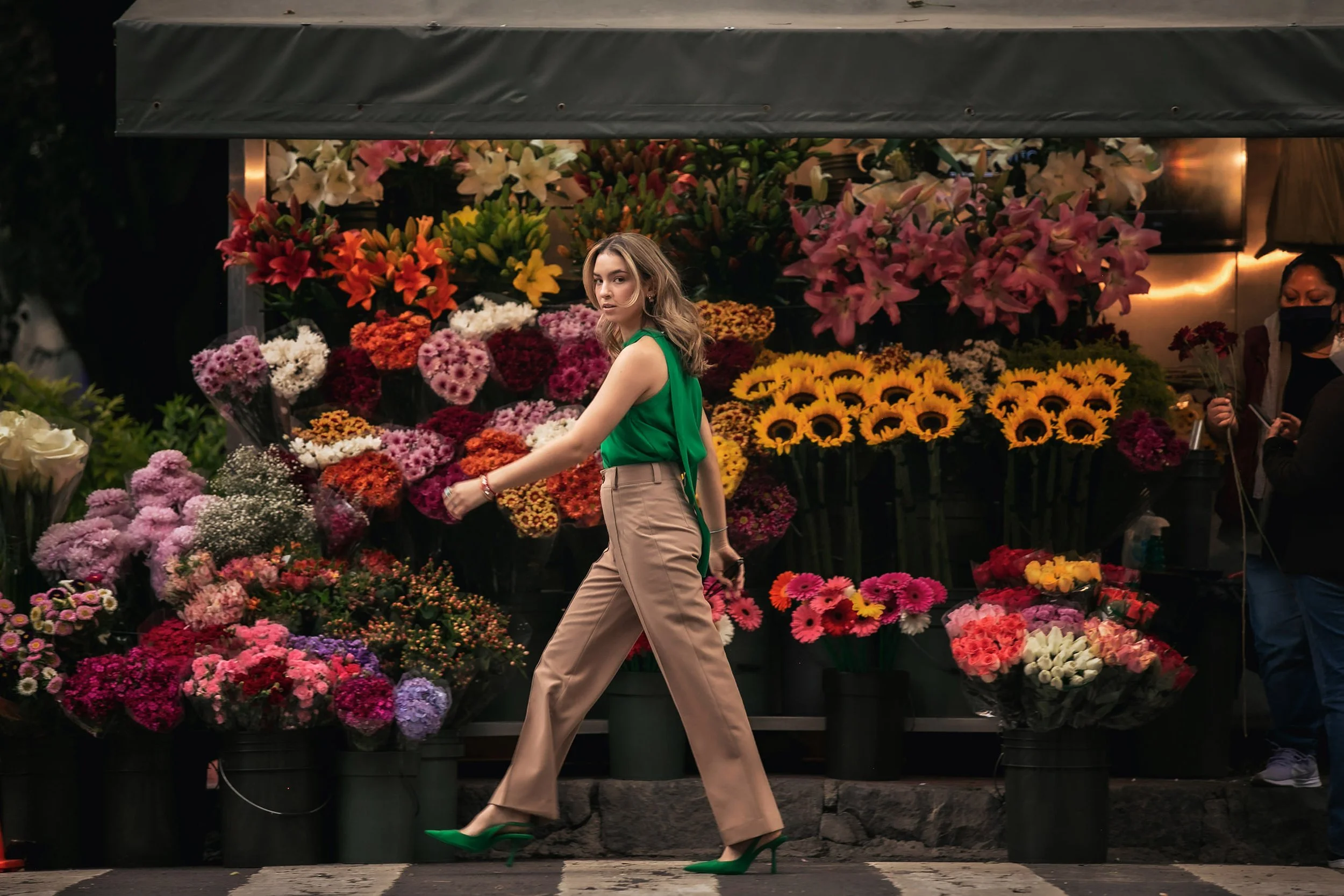 Retrato de mujer caminando frente a puestos de flores, escena urbana colorida.