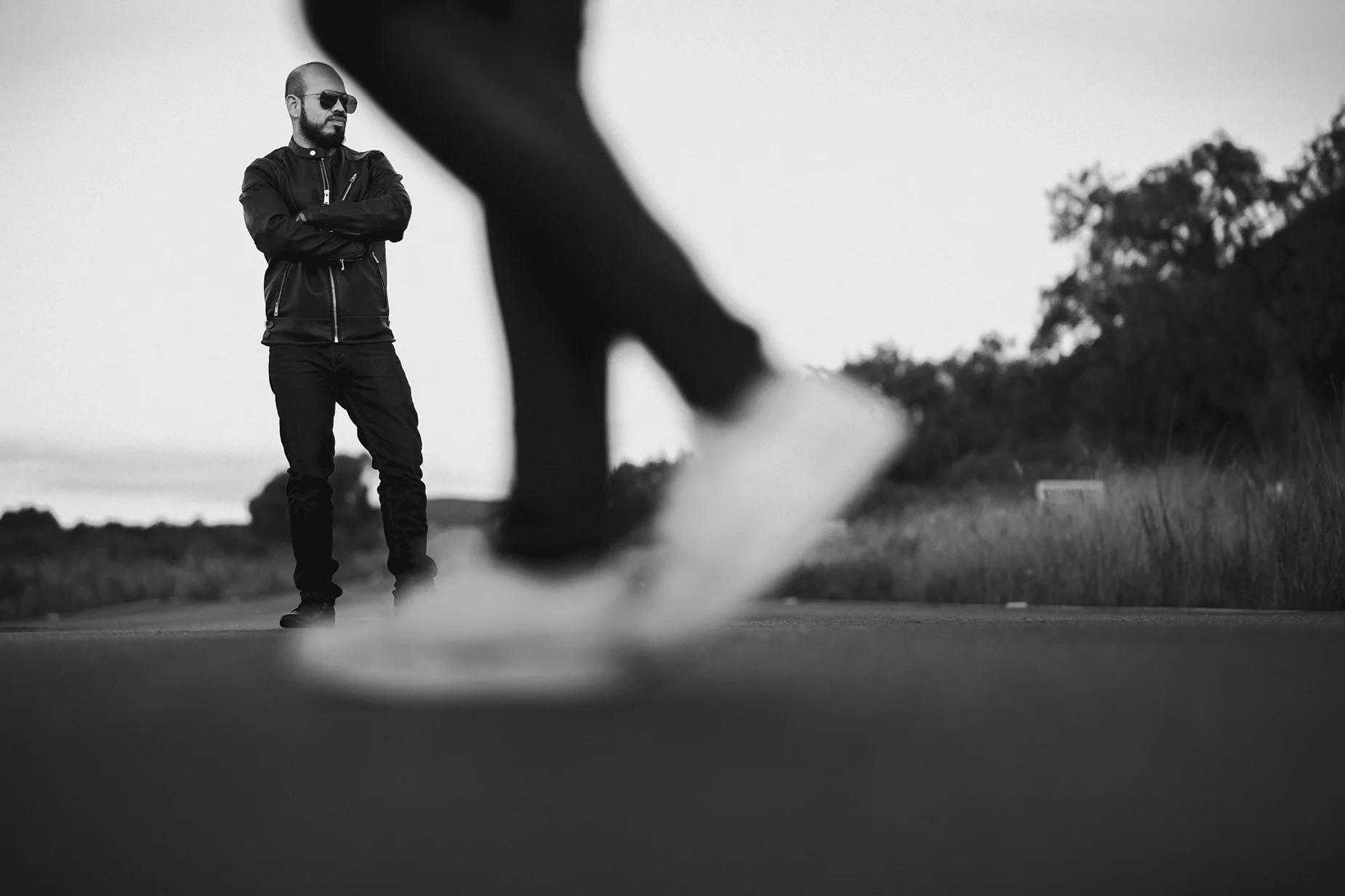 fotografia blanco y negro de hombre vestido de negro parado en la carretera con unas piernas de alguien caminando en primer plano.