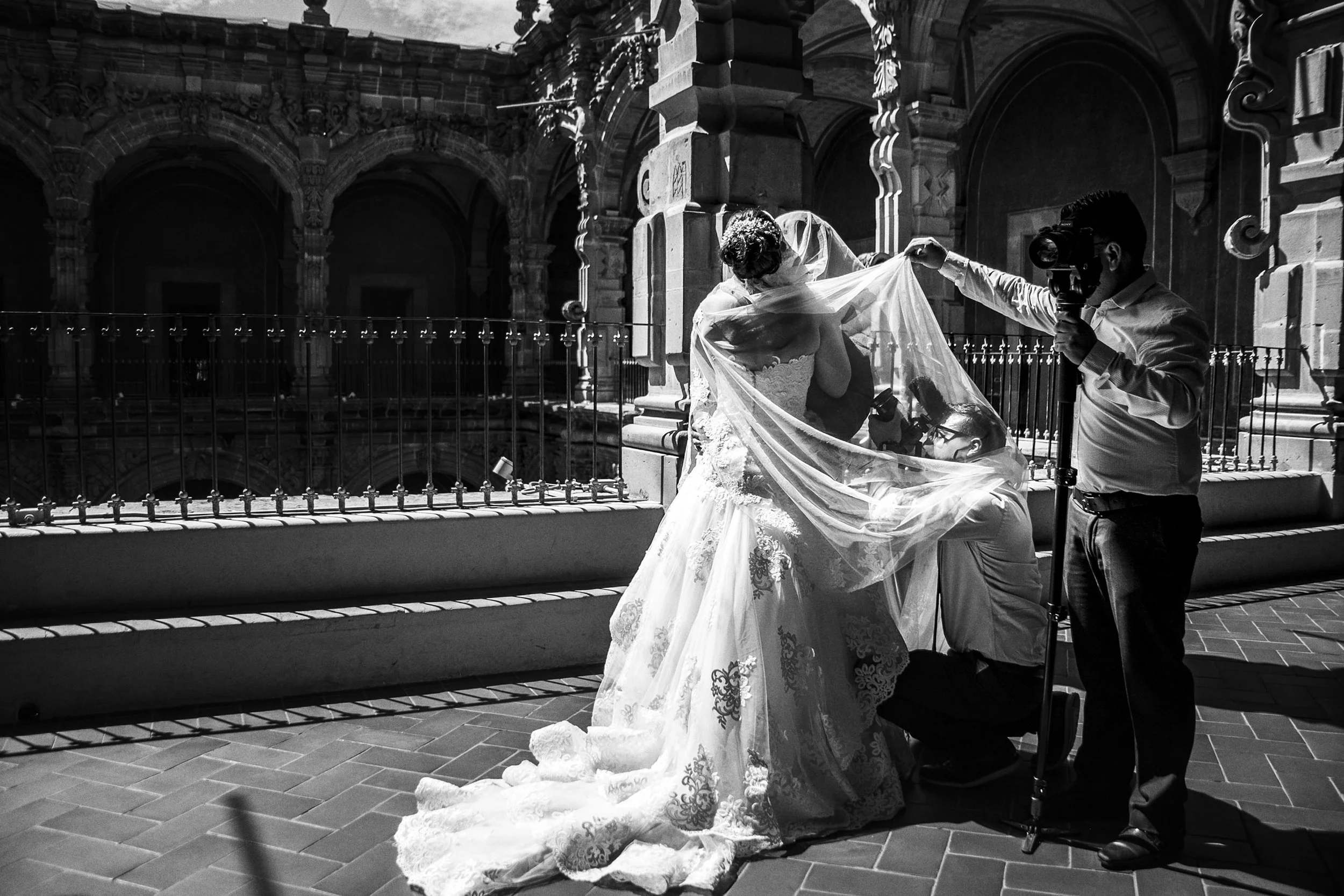 Fotografía en blanco y negro de novia ajustando vestido en exterior urbano, tela en movimiento.