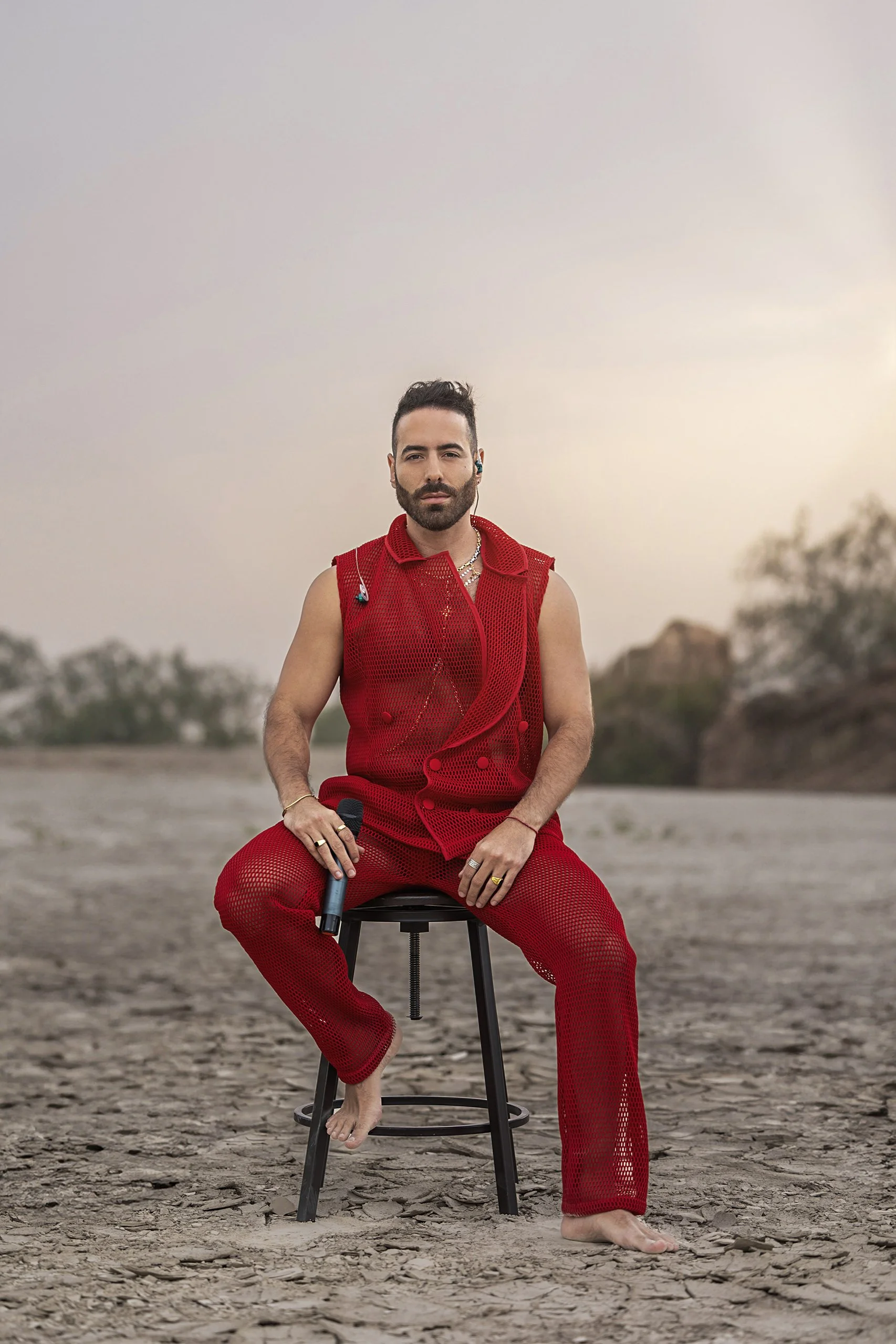 Retrato de hombre sentado en exterior desértico, vestimenta roja, postura firme y mirada directa.