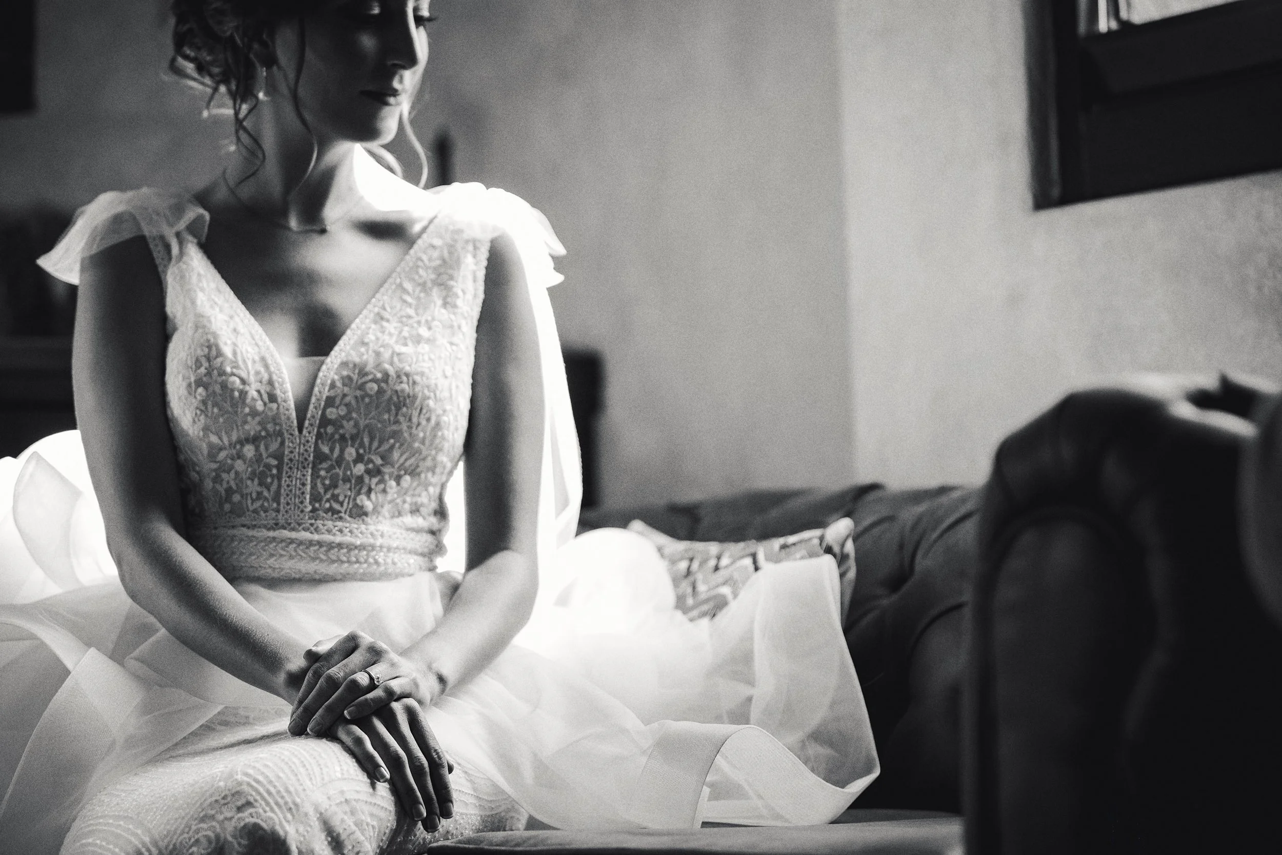 Fotografía en blanco y negro de novia sentada durante preparación, manos apoyadas sobre el vestido.