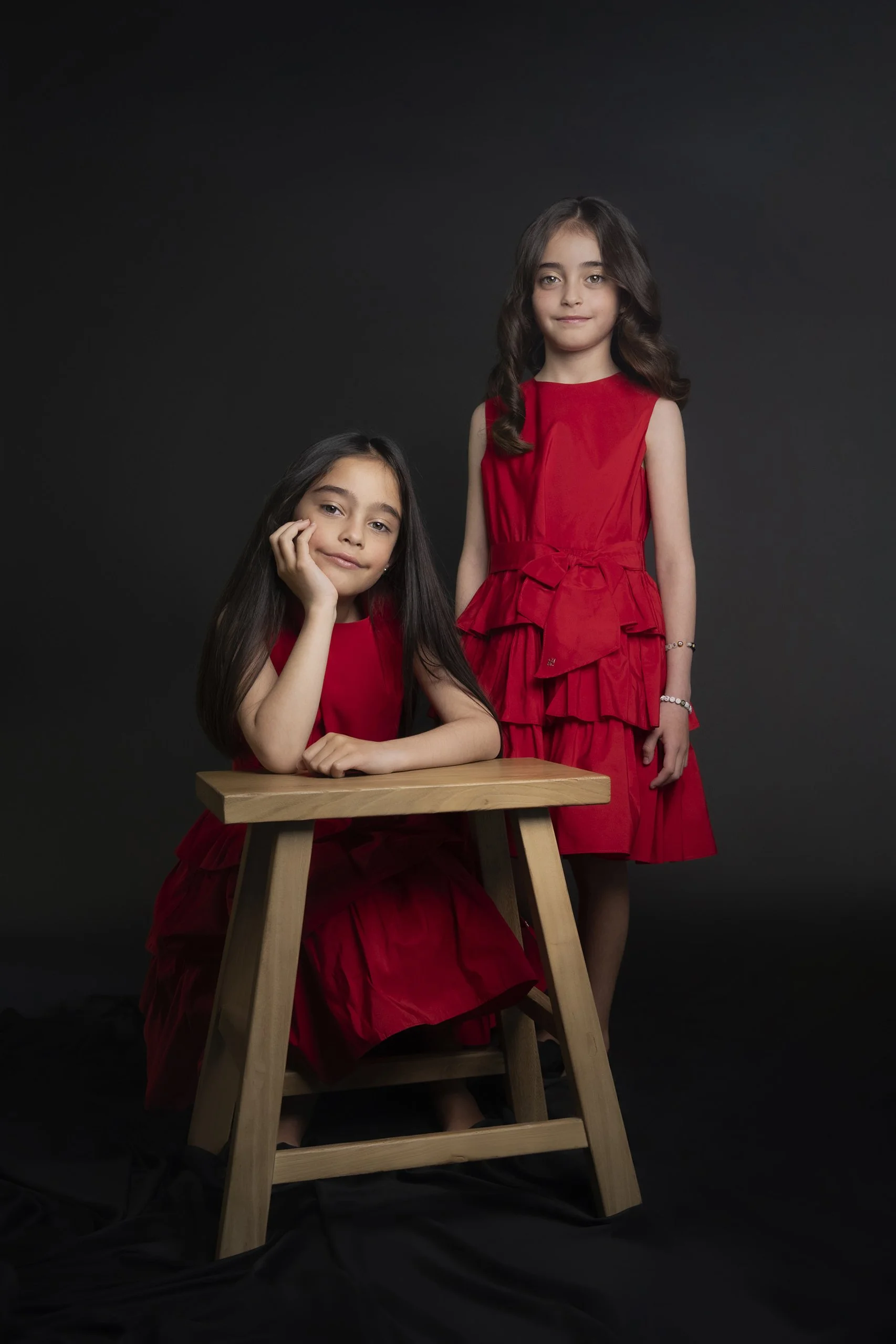 Retrato de dos niñas con vestidos rojos en estudio, una sentada y otra de pie, expresión serena y cercana.