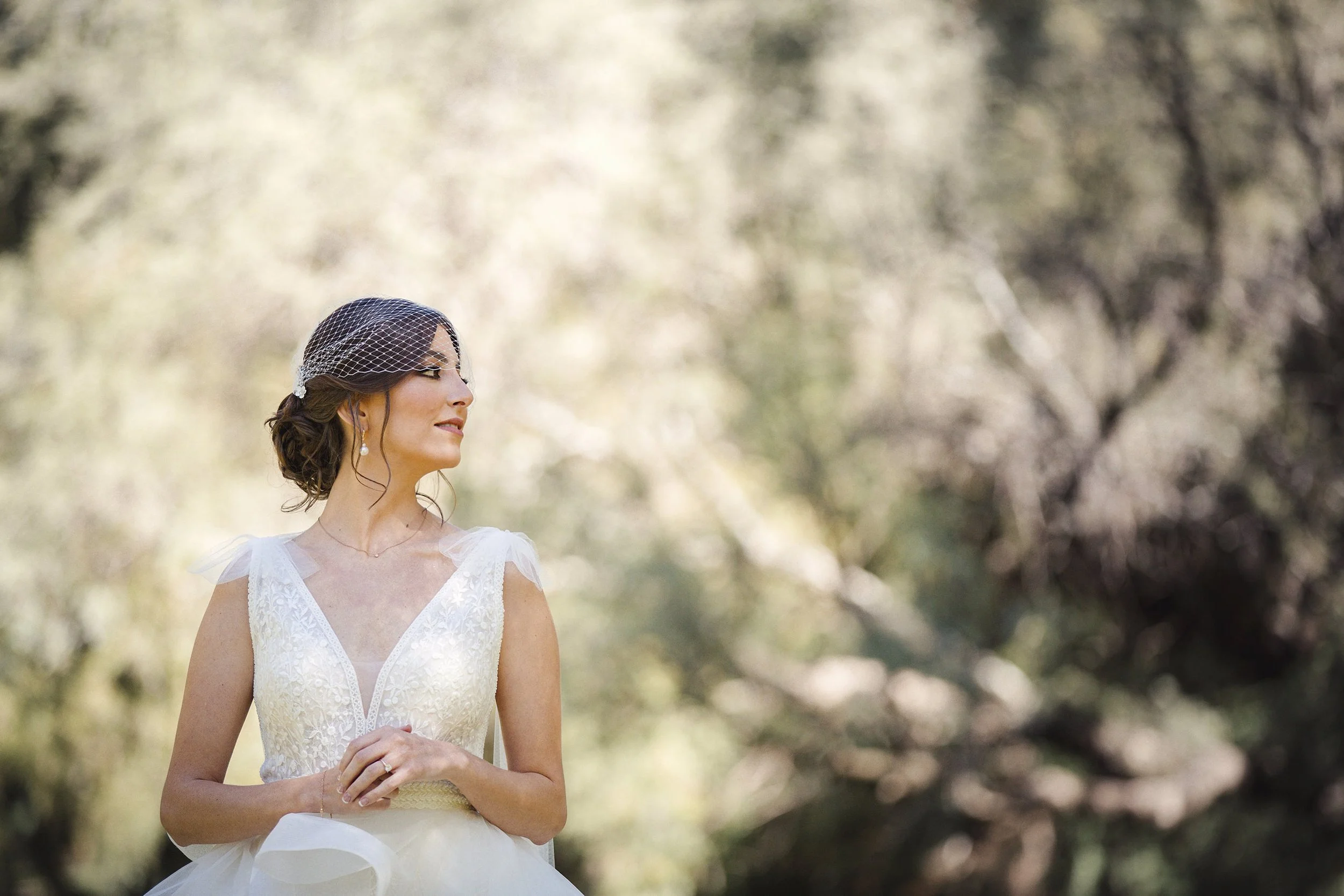 Fotografía de novia en exterior, vestida de blanco y mirando hacia un lado entre árboles.