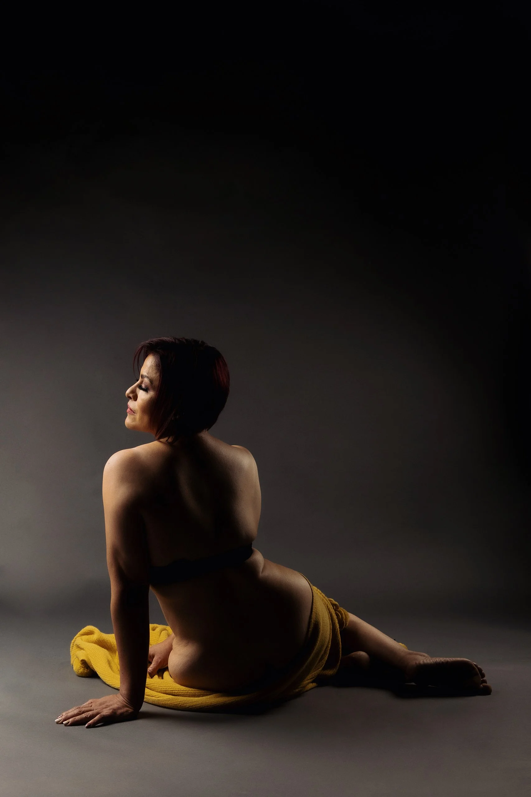 Retrato de mujer sentada de perfil en estudio, espalda descubierta y luz suave sobre fondo oscuro.
