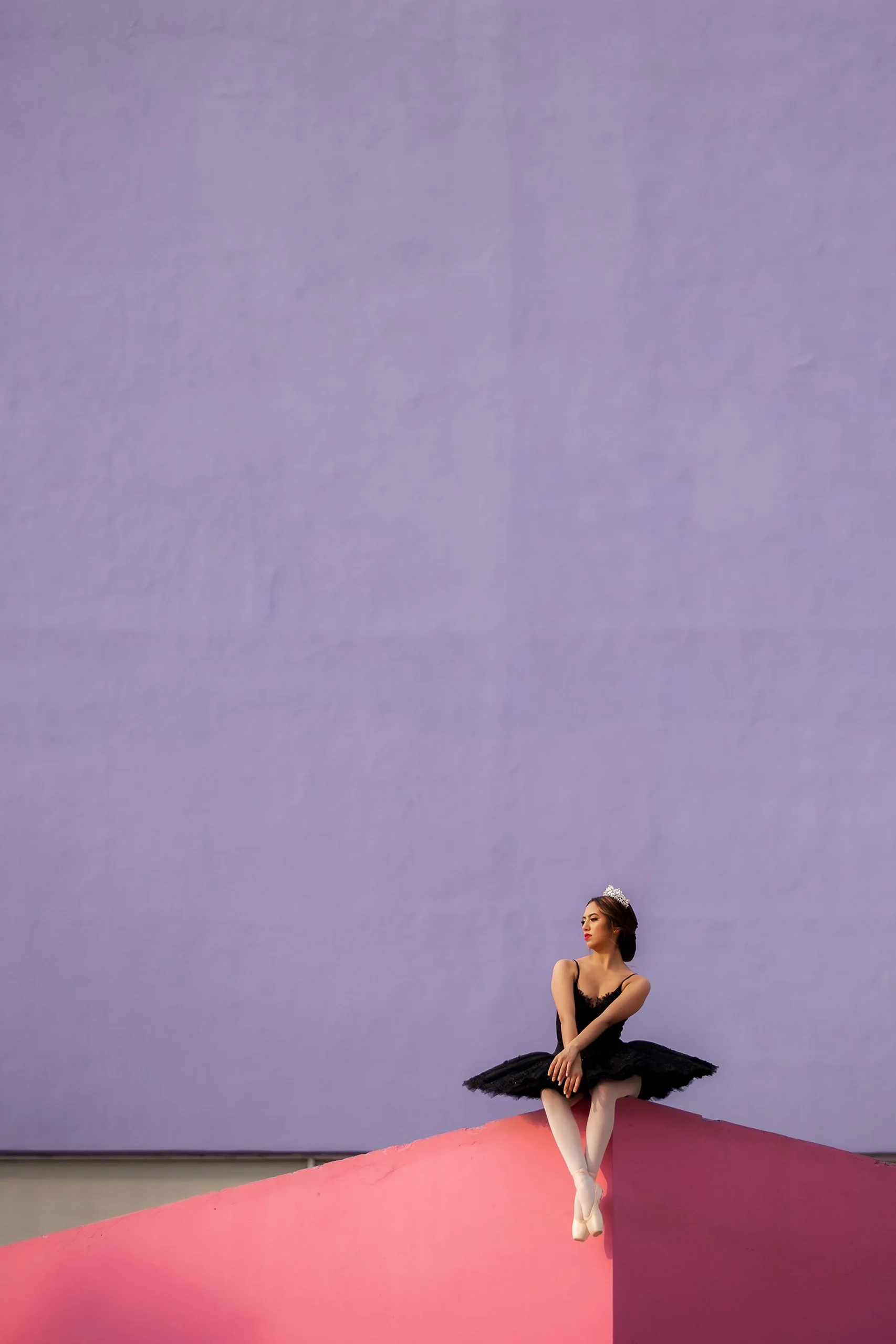 Retrato de mujer sentada en exterior minimalista frente a fondo liso color violeta, figura pequeña y postura tranquila.