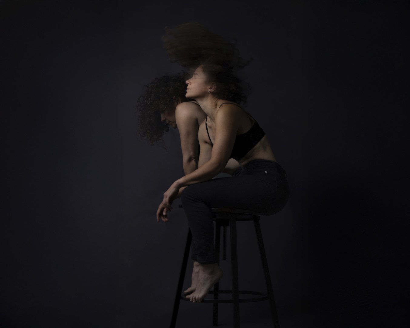 A woman with curly hair sitting on a black stool against a dark background, captured in multiple overlapping motion blur images, wearing a black tank top and jeans.