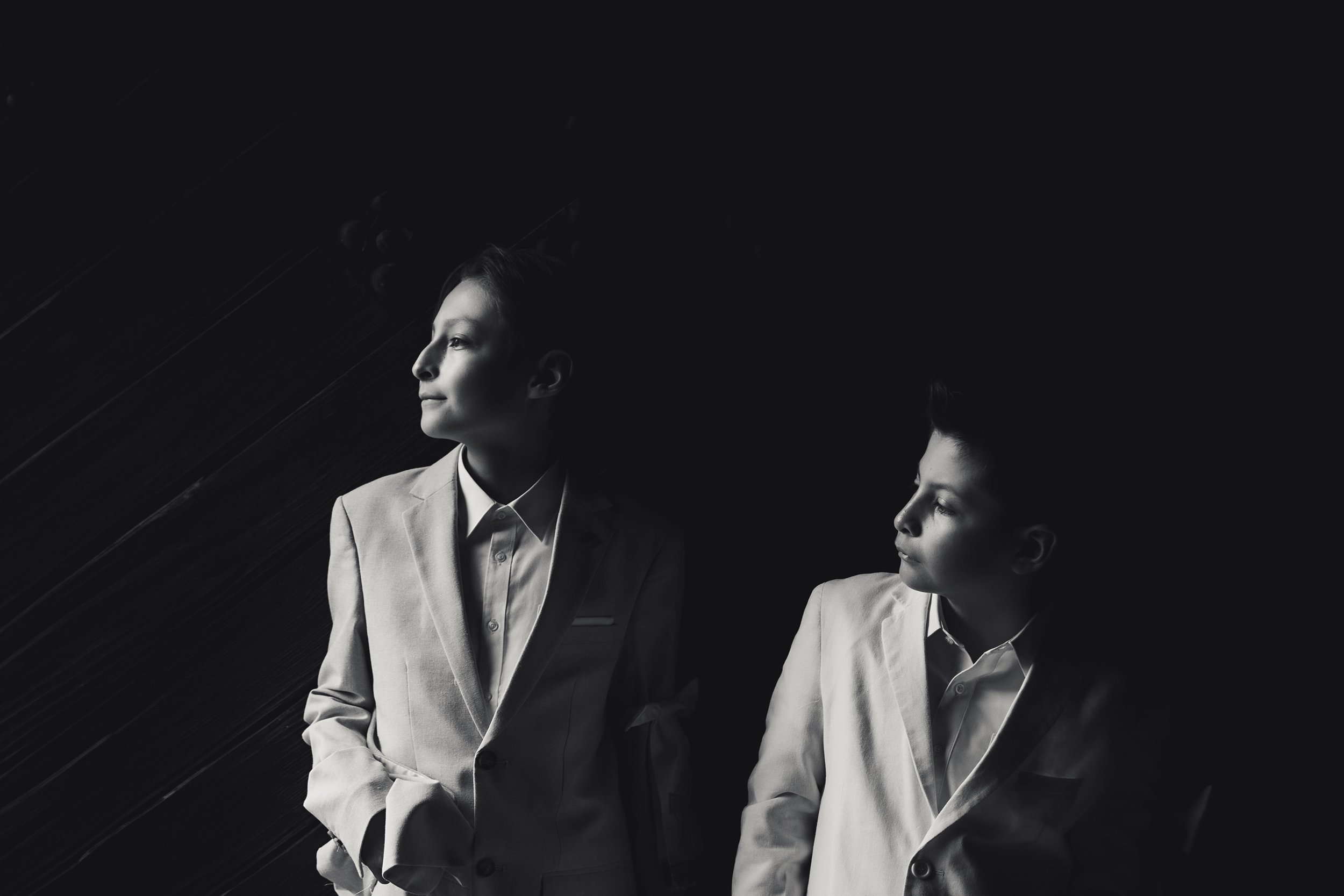 Retrato en blanco y negro de dos personas con traje formal, de pie y mirando hacia el mismo punto, fondo oscuro.