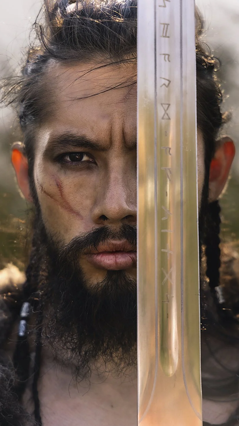 Close-up of a man with long hair and beard, holding a sword vertically in front of his face, with part of his face showing on the left side.