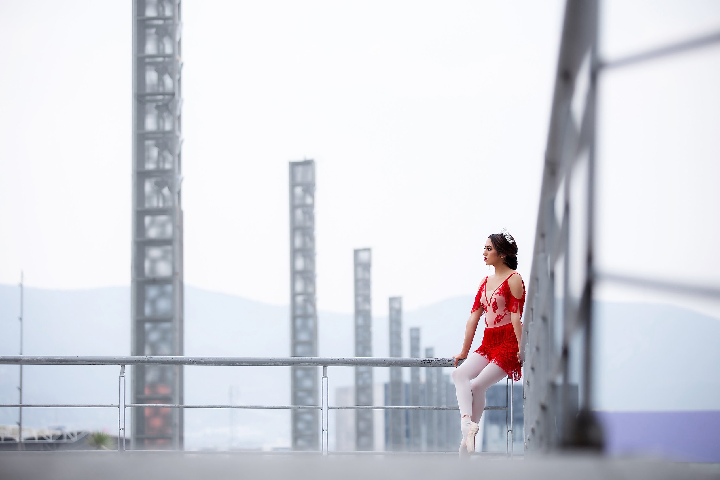 Retrato de niña con vestido rojo sentada junto a barandal metálico, escena urbana con fondo arquitectónico.