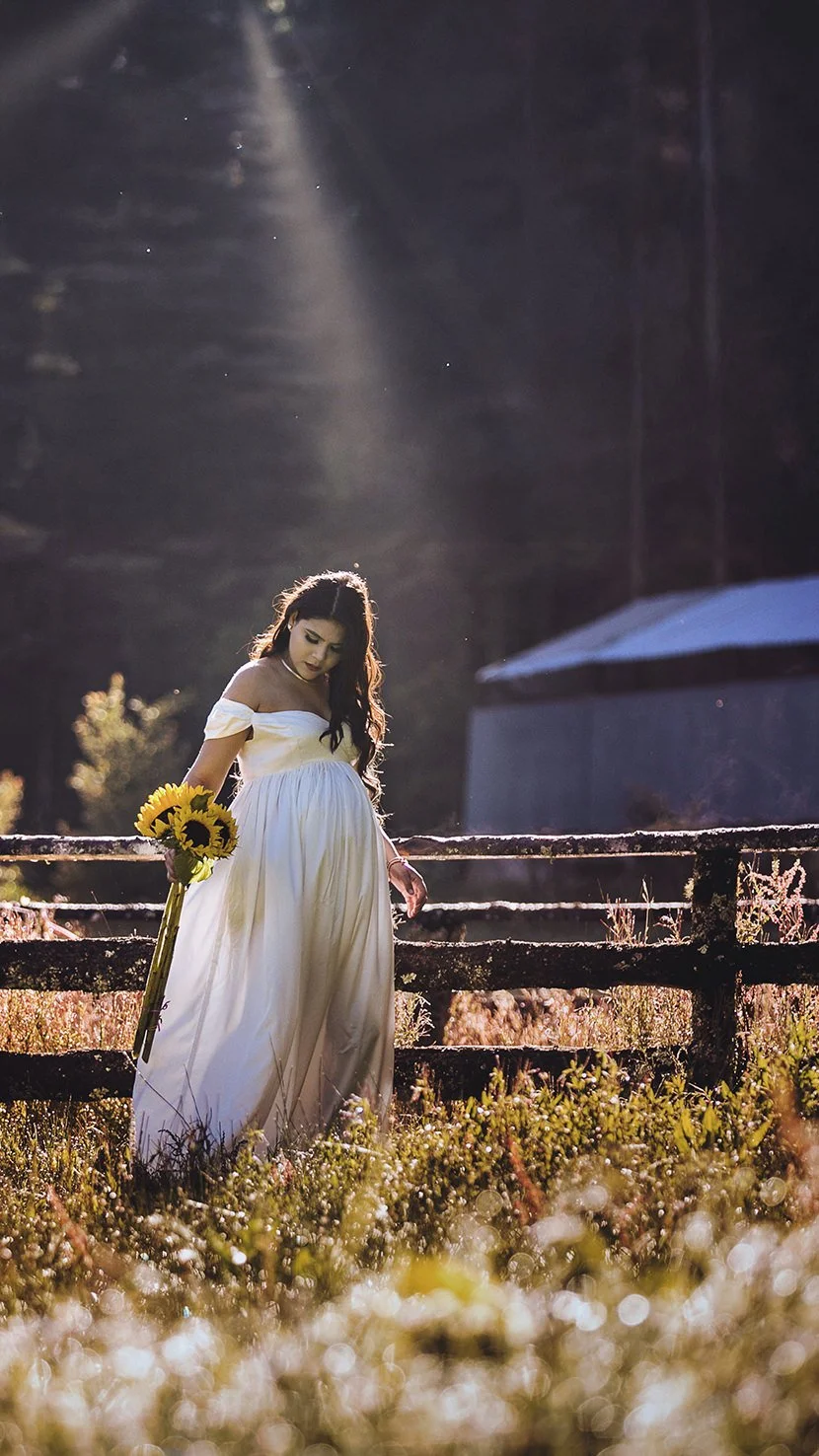 Pregnant woman in a white dress holding a bouquet of sunflowers stands outdoors near a rustic wooden fence at sunset.