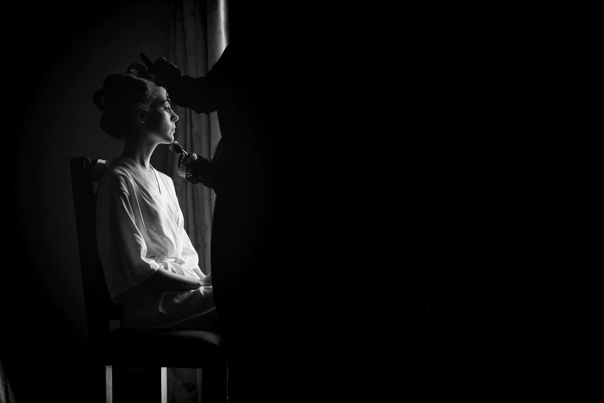 Fotografía en blanco y negro de novia sentada junto a ventana, luz lateral y gesto reflexivo.