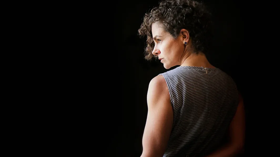 Side profile of a woman with curly hair looking to her left, wearing a sleeveless striped shirt, against a dark background.
