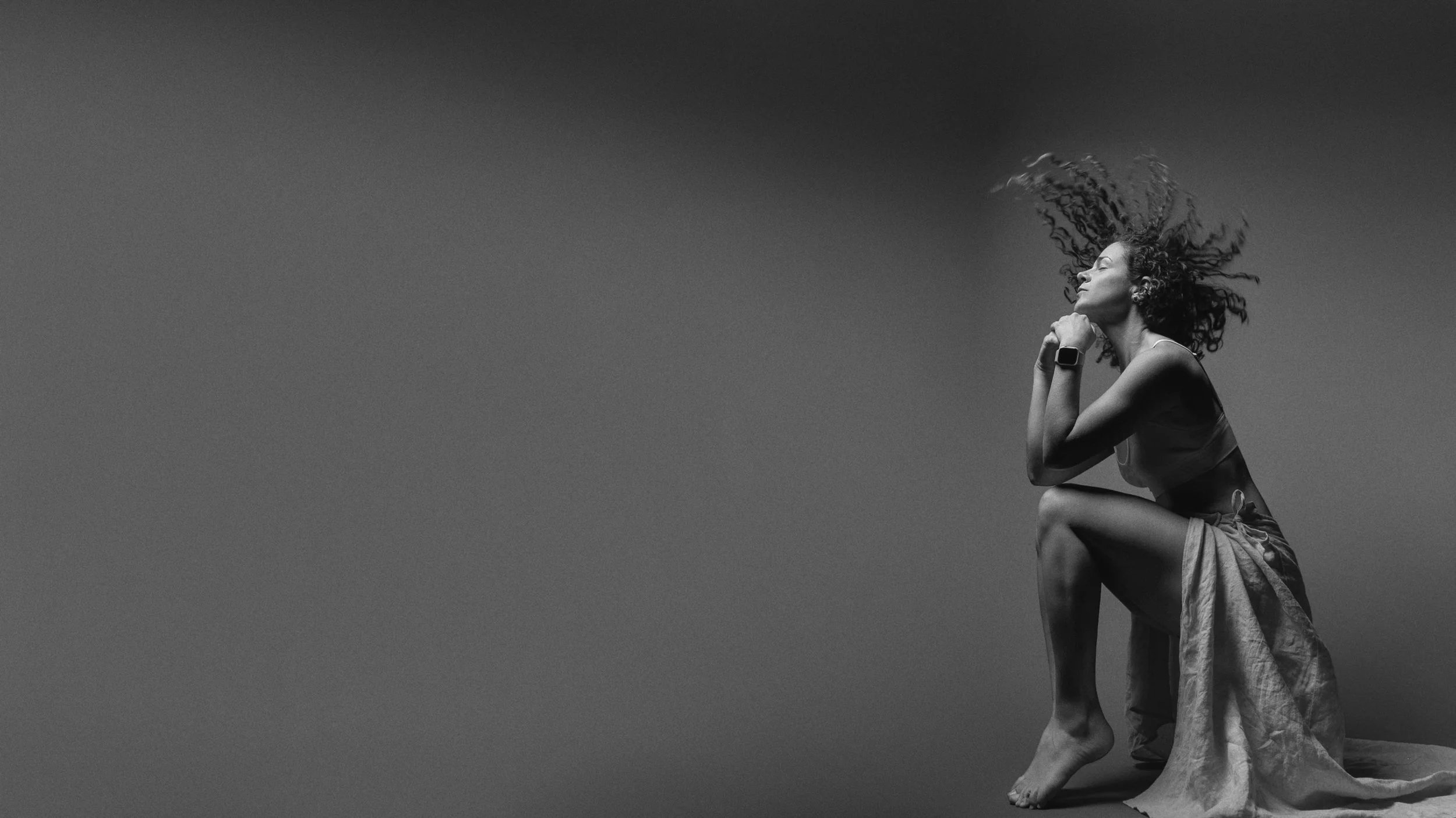 A woman with curly hair sitting barefoot on the ground, in profile, with her eyes closed and head tilted back, creating a sense of serenity and contemplation.