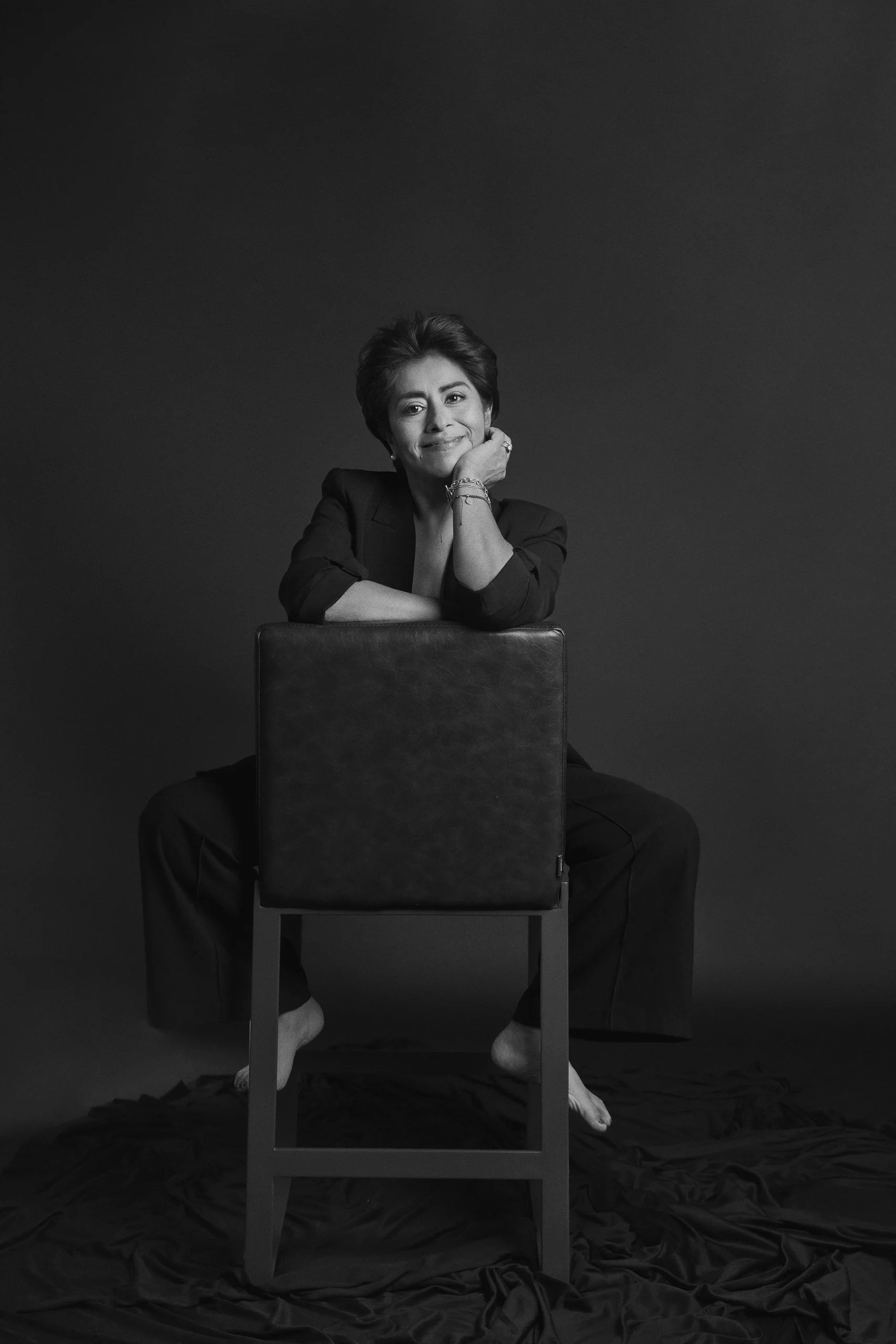 Retrato en blanco y negro de mujer sentada, mirada directa y gesto reflexivo, fondo oscuro.