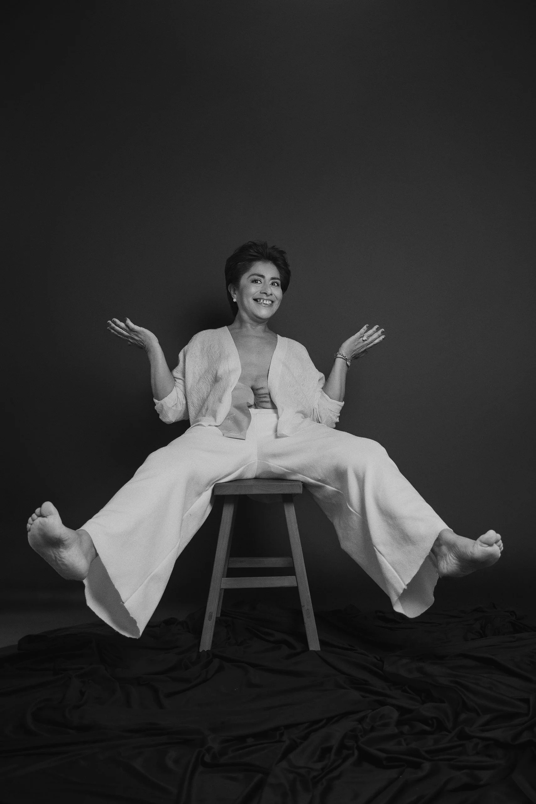 Retrato en blanco y negro de mujer sentada con brazos abiertos y gesto espontáneo, fondo oscuro.