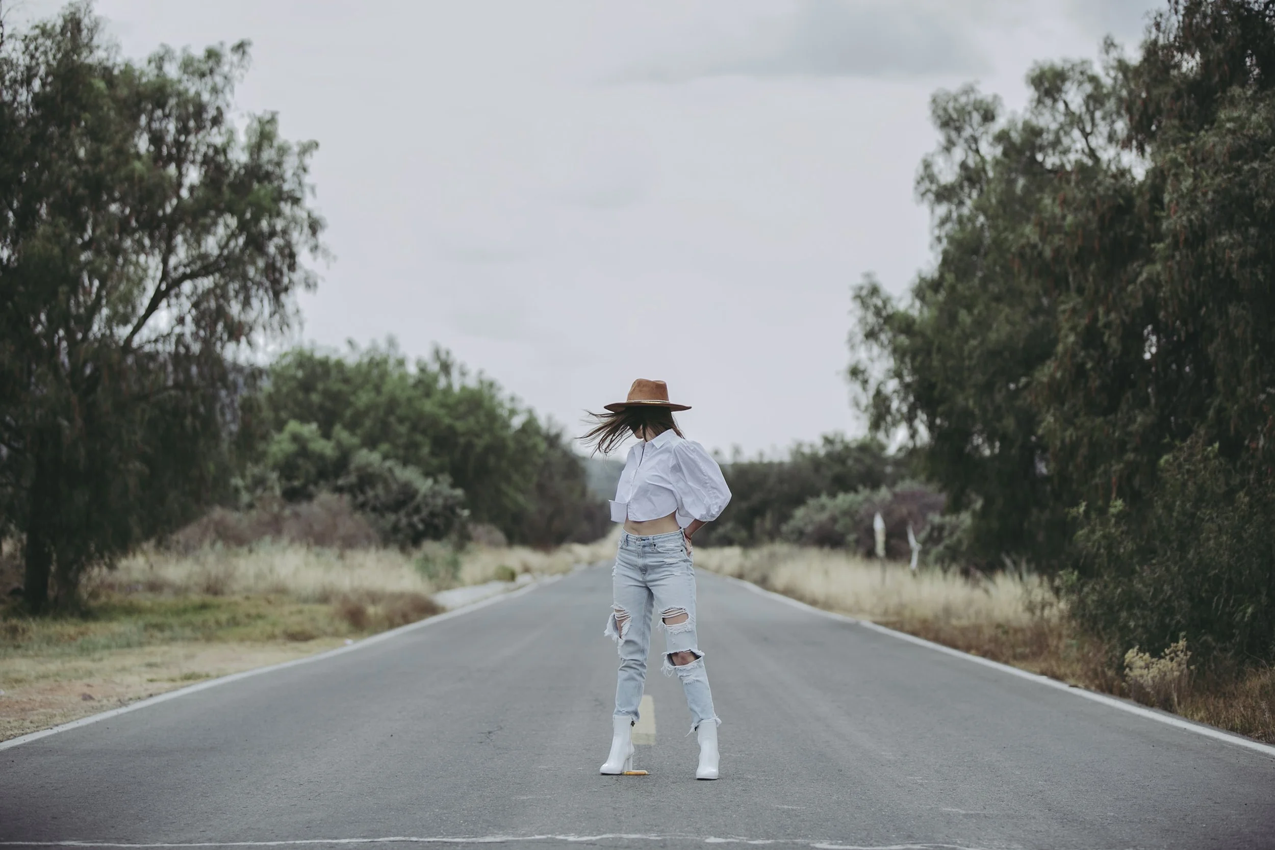 Retrato de mujer de espaldas caminando por carretera rural, vestimenta clara y sombrero, escena en exterior.