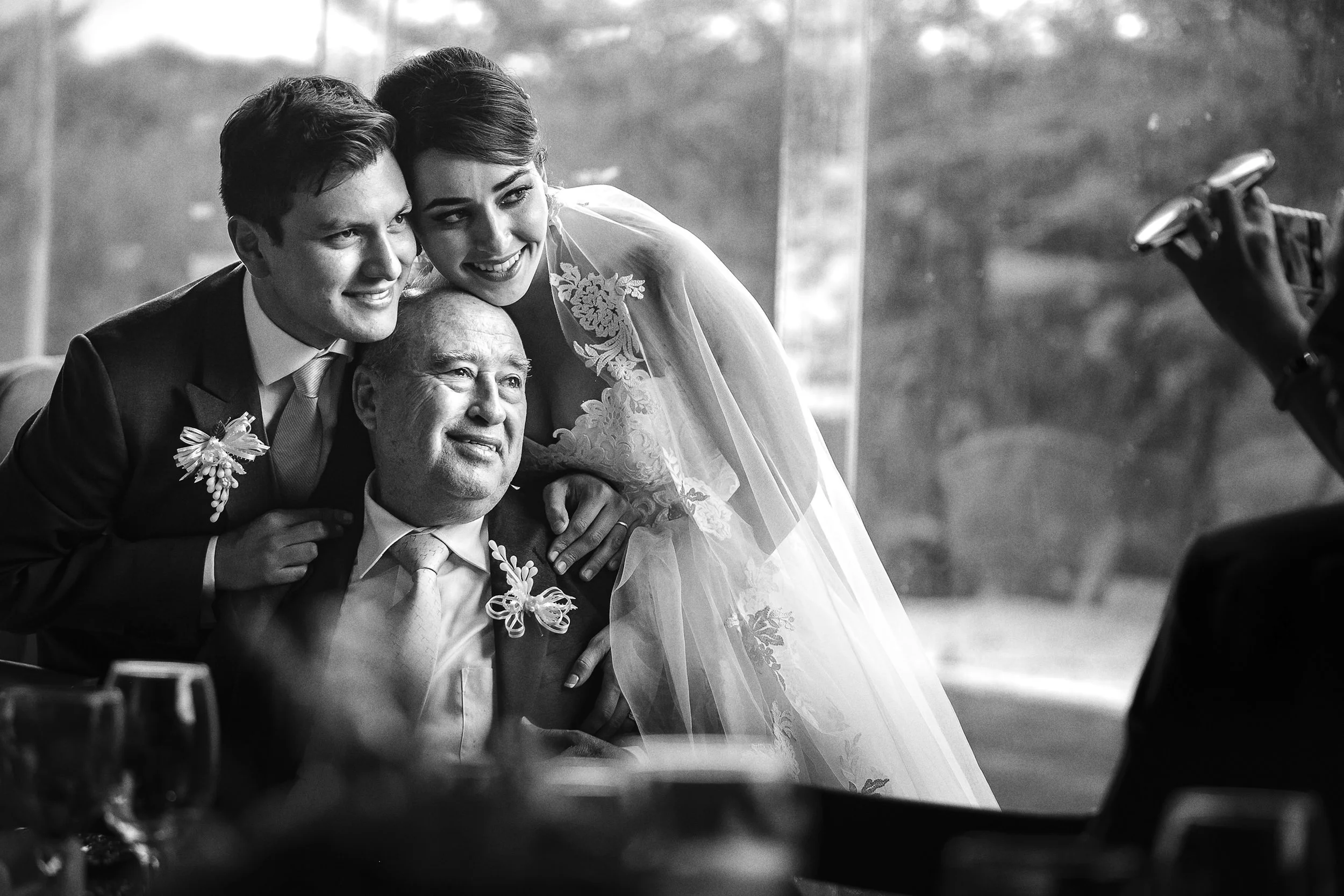 Fotografía en blanco y negro de novios acompañados por familiar mayor, gesto cercano y emotivo.