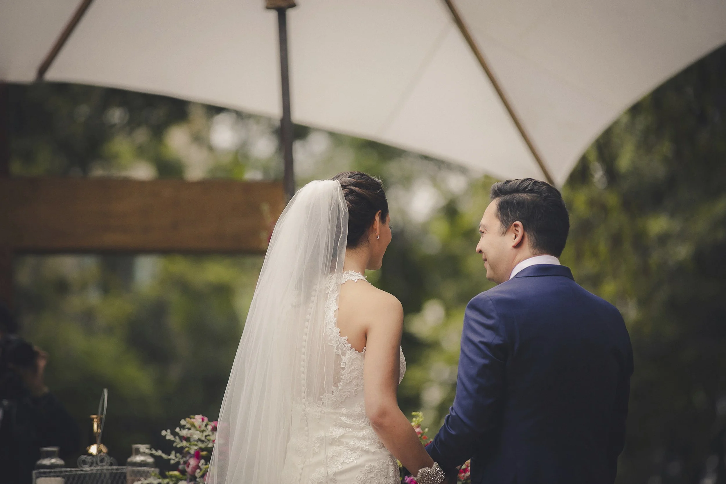 Fotografía de novios de espaldas tomados de la mano durante ceremonia al aire libre.